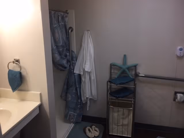 A bathroom with a sink and countertop on the left, a shower with a blue patterned curtain, a white robe hanging on a hook, a metal rack holding folded towels and a starfish decoration, a grab bar on the wall, and a pair of slippers on the floor.