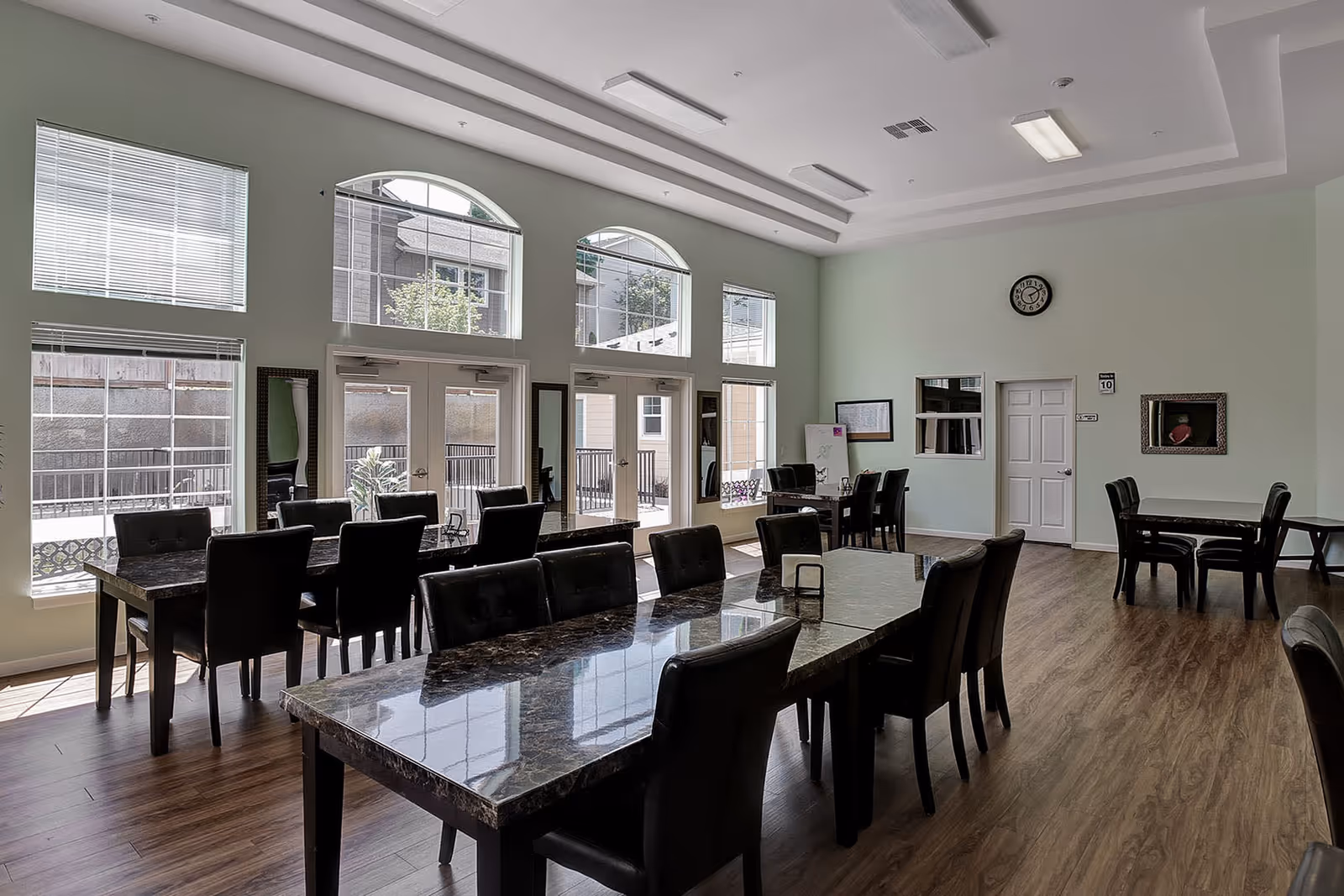 A spacious dining room with multiple dark marble tables and black chairs arranged neatly. Large windows and glass doors allow natural light to fill the room. The walls are light-colored, and there is a clock and framed items on the far wall. The floor is wooden, and the ceiling has recessed lighting.