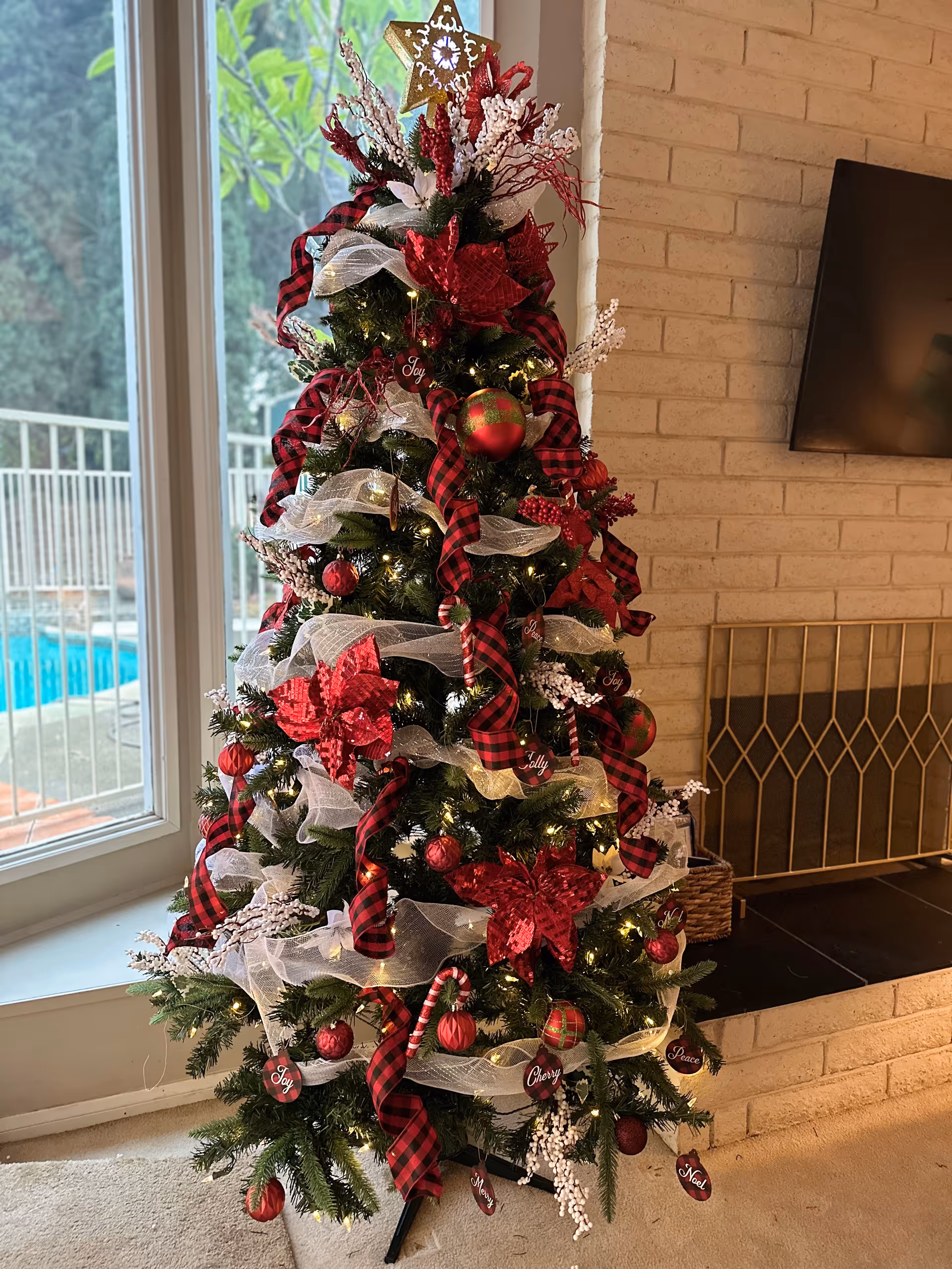Decorated Christmas tree with red and white ribbons and ornaments next to a fireplace and window.