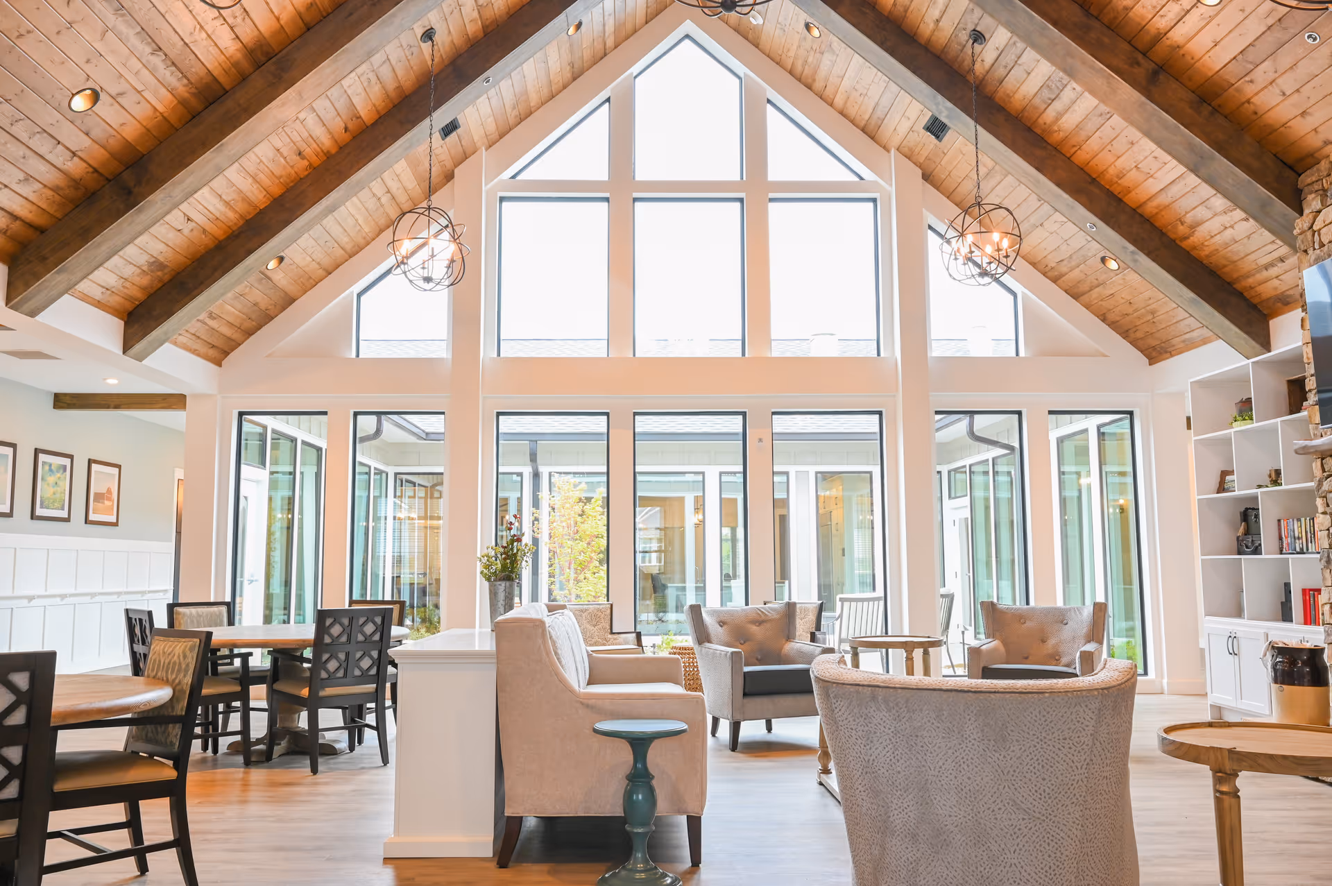 A bright and spacious senior living common area with high vaulted wooden ceilings and large triangular windows letting in natural light. The room features comfortable armchairs arranged around small tables, dining tables with chairs, and built-in shelves with books and decor. The space has a warm and inviting atmosphere with light-colored walls and wooden flooring.