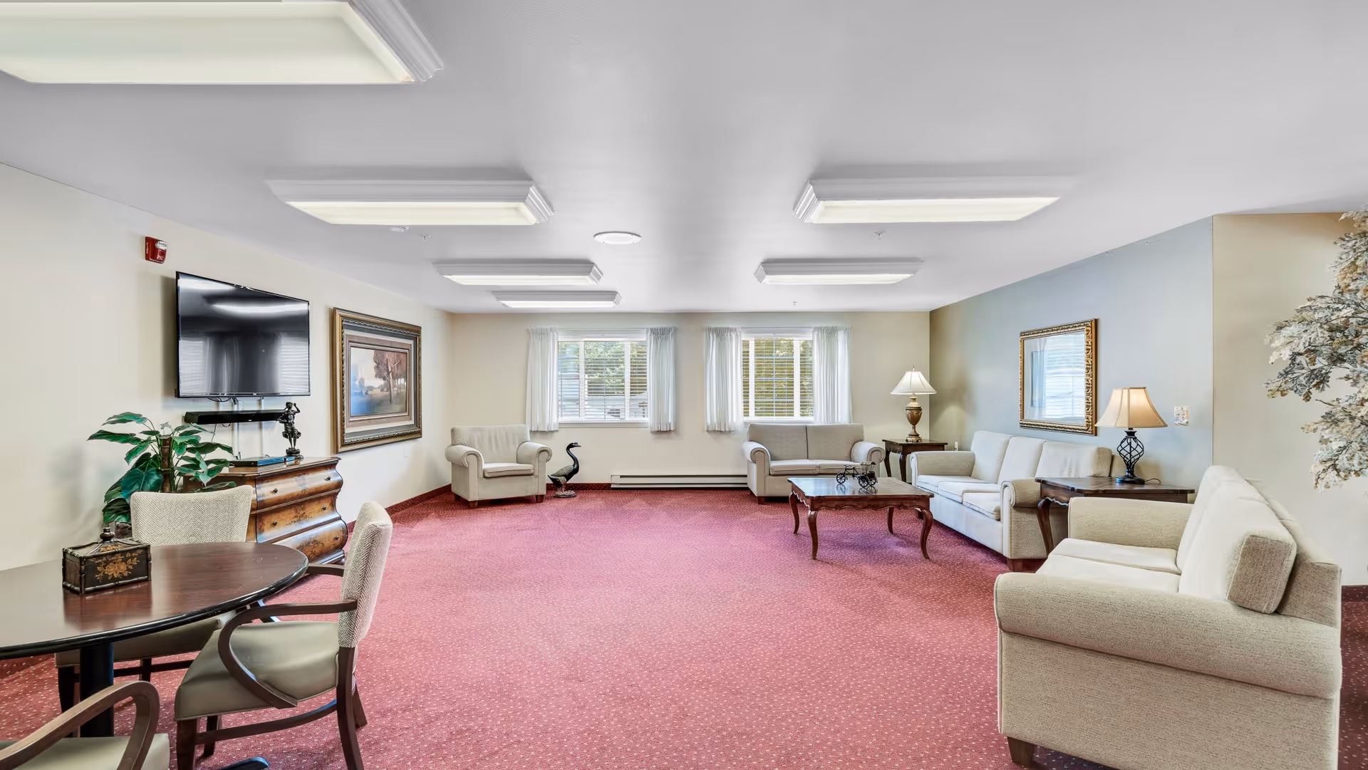 Spacious communal living room with sofas, armchairs, coffee tables, a wall-mounted TV, and red carpeting.
