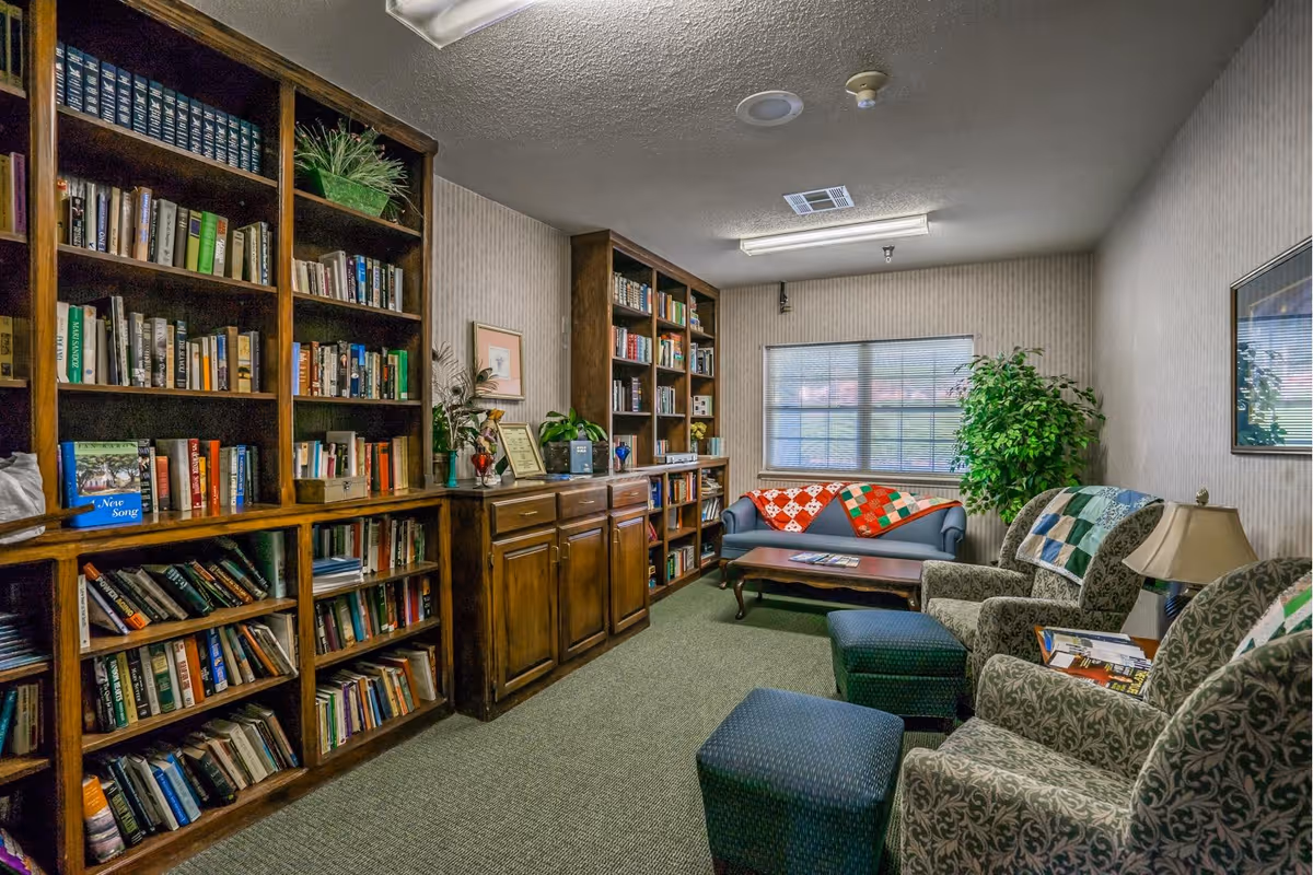 A cozy reading room with wooden bookshelves filled with books along the left wall. There are two patterned armchairs with ottomans, a small table lamp, and a coffee table in front of a blue sofa adorned with colorful quilts. A large window with blinds and a green potted plant are on the far wall.