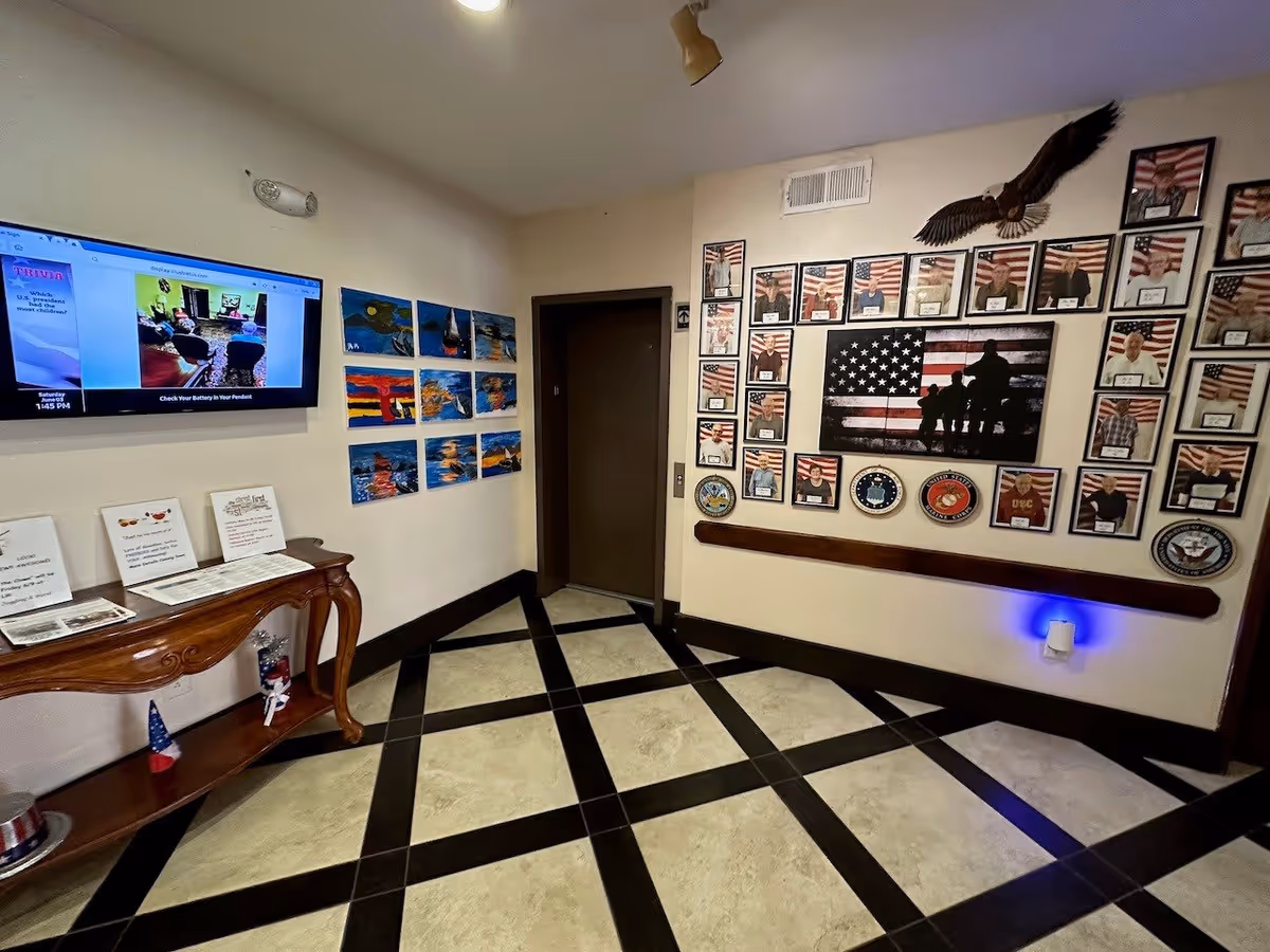 Interior corner of a senior living facility hallway with a wall-mounted TV displaying trivia, a wooden table with informational signs, and a wall decorated with framed photos of veterans, military emblems, and an American flag artwork. The floor has a beige and black checkered tile pattern.