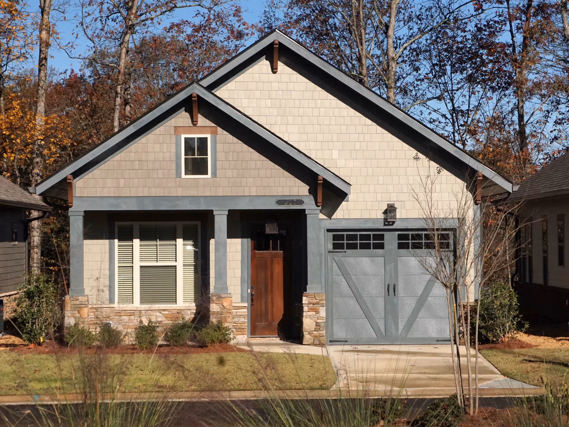 Front exterior view of a single-story house with beige siding, a wooden front door, a garage with gray doors, and a small front yard with grass and shrubs. Trees with autumn leaves are visible in the background.