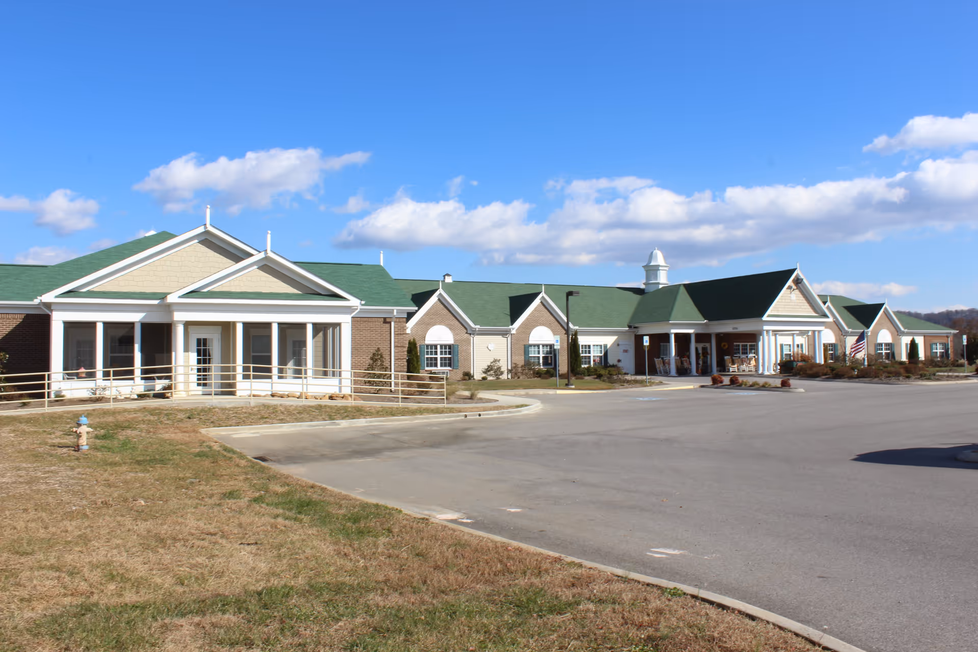 Exterior front view of a single-story senior living facility with green roofs, a driveway and clear blue sky.