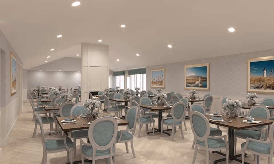 A spacious dining room with multiple wooden tables and light blue upholstered chairs arranged neatly. Each table is set with plates, glasses, and floral centerpieces. The room has soft lighting from recessed ceiling lights and framed artwork on the walls depicting serene outdoor scenes.
