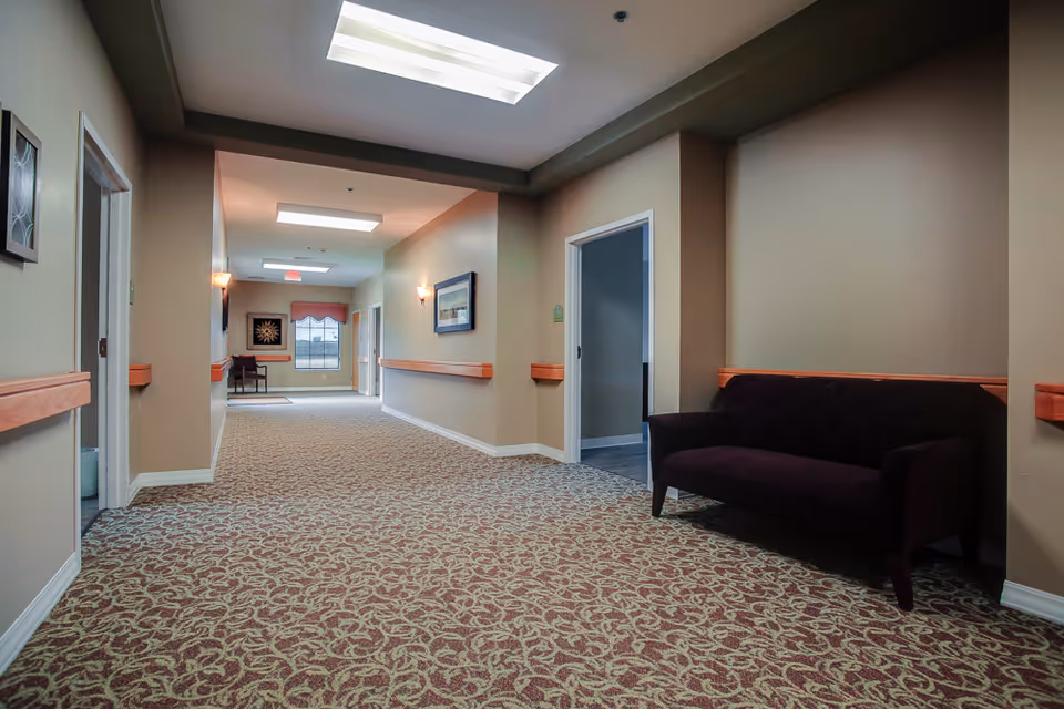 A well-lit hallway in a senior living facility with patterned carpet, beige walls, and wooden handrails. There is a dark purple upholstered bench on the right side and a framed picture on the wall. At the end of the hallway, there is a window with a valance and a small table with a chair underneath it.