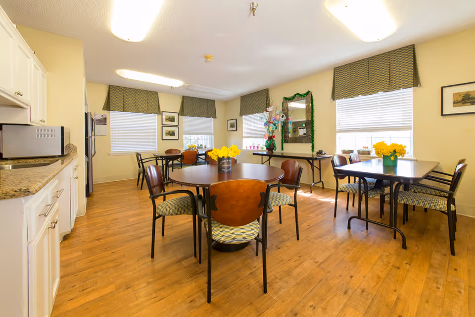 A bright dining room with wooden floors, multiple tables and chairs, and yellow flowers in vases on the tables. The room has three windows with green valances, a large mirror decorated with green garland, and a kitchenette area with white cabinets and a microwave.