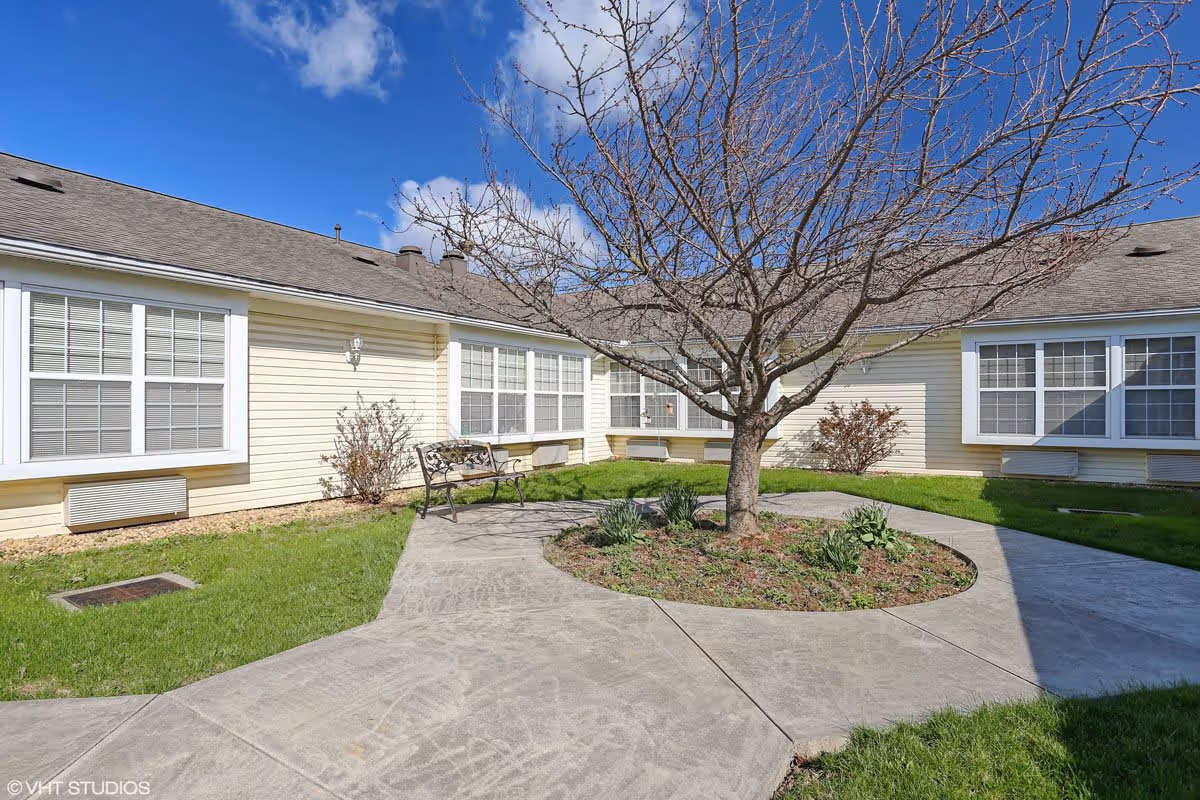 Outdoor courtyard area of a senior living facility with a leafless tree in the center surrounded by a circular garden bed. There is a concrete walkway around the tree and a metal bench on the left side. The building has light yellow siding with multiple large windows and a gray shingled roof under a blue sky with some clouds.