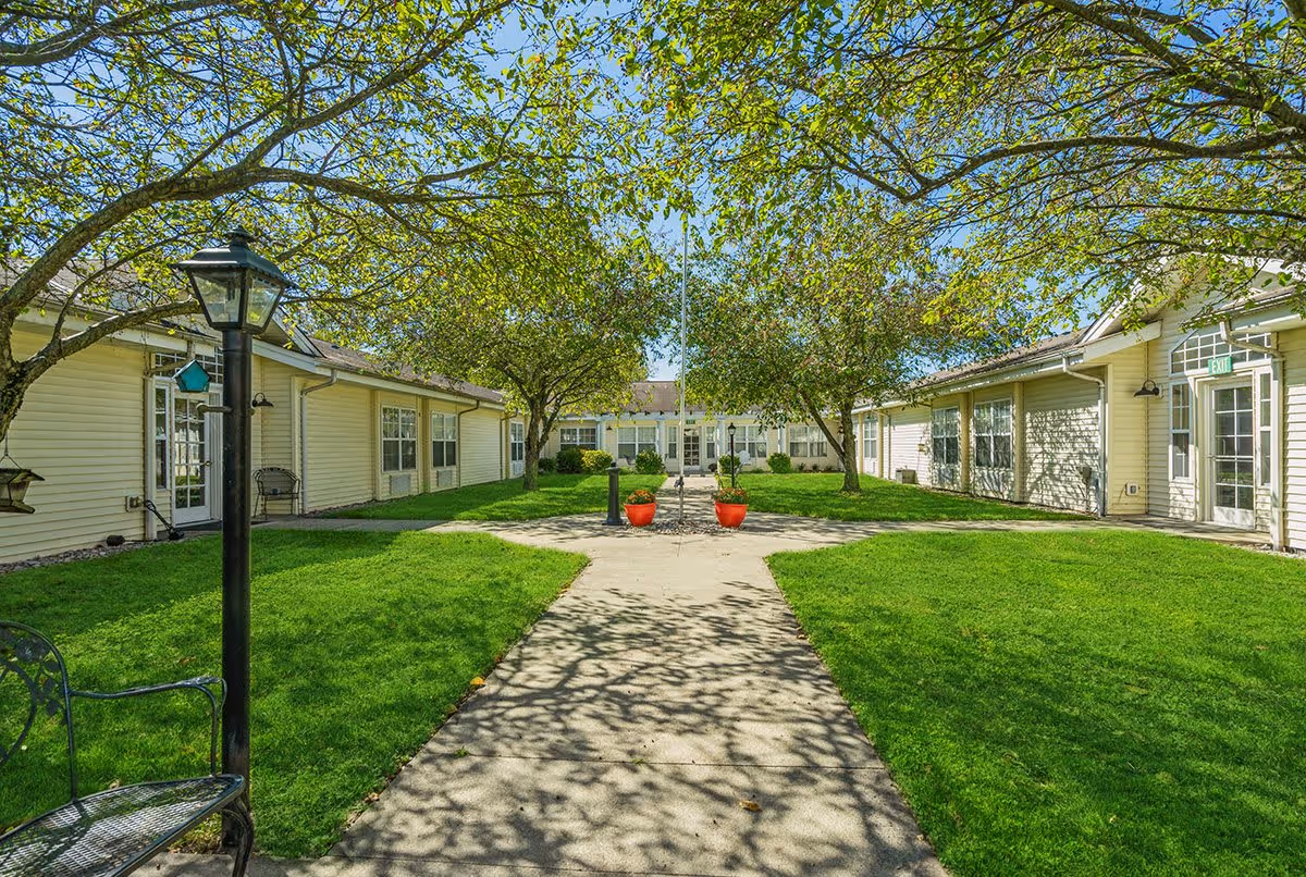 A sunny courtyard area with a concrete pathway leading through green grass and trees with light green leaves. The courtyard is surrounded by single-story beige buildings with multiple windows and glass doors. There are two red flower pots on either side of the pathway near the center, and a black lamp post and metal bench are visible on the left side.