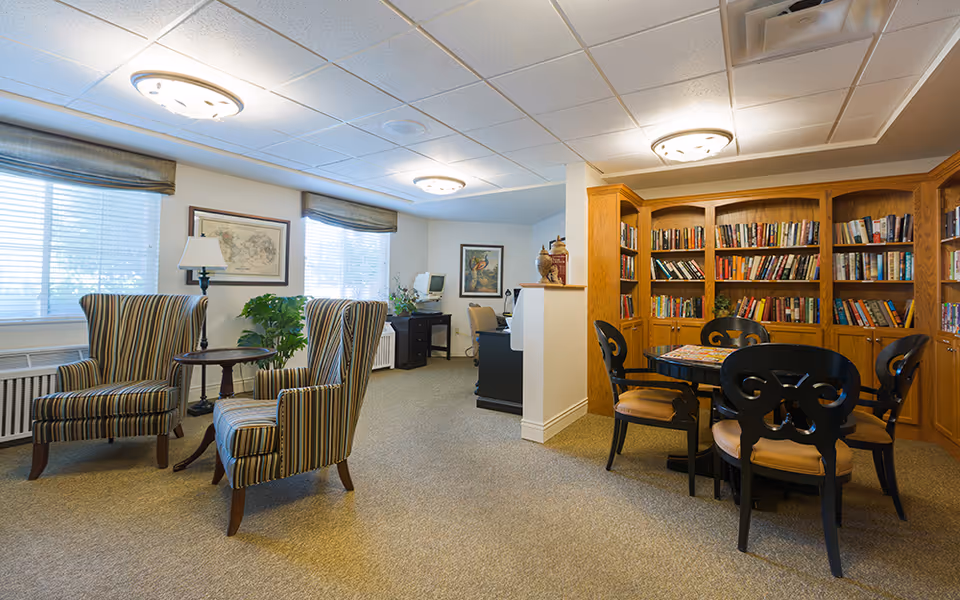 A cozy senior living common area with two striped armchairs and a small round wooden table near two windows with blinds and valances. On the right side, there is a wooden bookshelf filled with books and a round table with four black chairs. The room has beige carpet, white walls, and ceiling lights. A desk with a computer and chair is visible in the background.