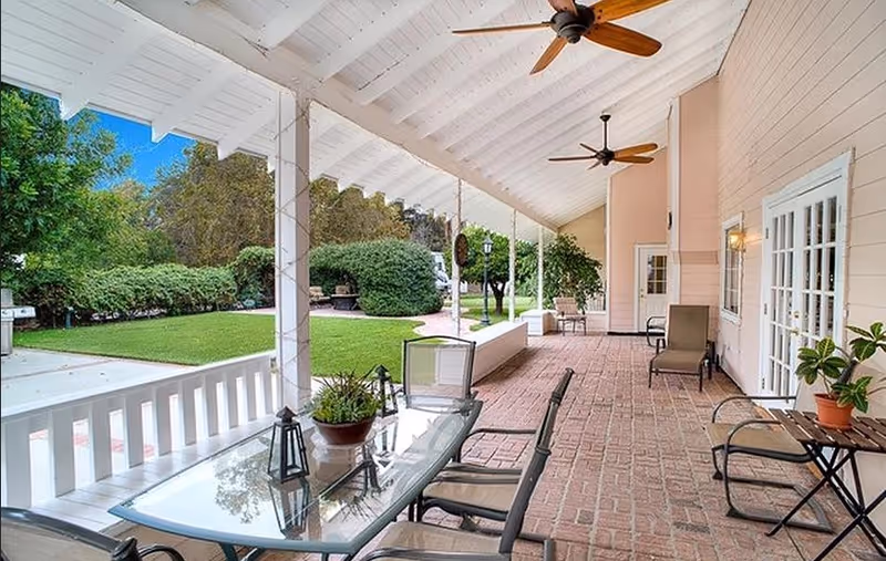 Covered patio with a glass dining table, chairs, ceiling fans, and potted plants overlooking a manicured lawn and garden.