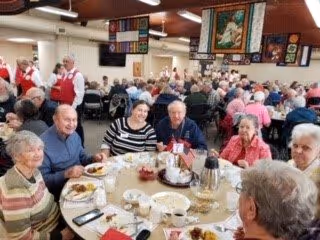 A large group of senior adults seated at round tables in a spacious dining room, enjoying a meal together. The room is decorated with colorful quilt-like wall hangings and has a warm, communal atmosphere.