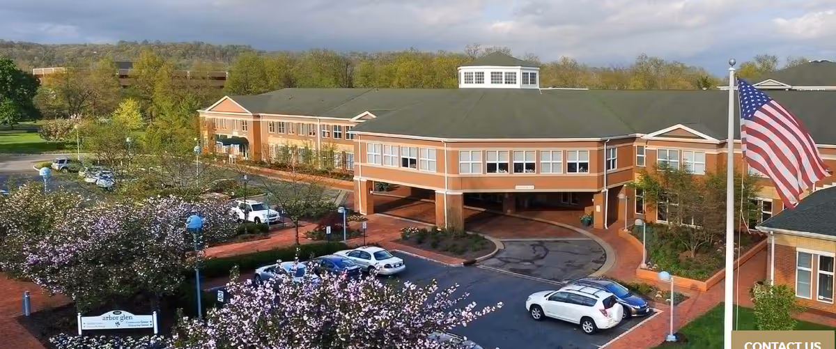 Aerial view of a senior living facility building with a large covered entrance, surrounded by parking spaces and blooming trees. An American flag is visible on a flagpole to the right, and the building is set against a backdrop of green trees and a partly cloudy sky.