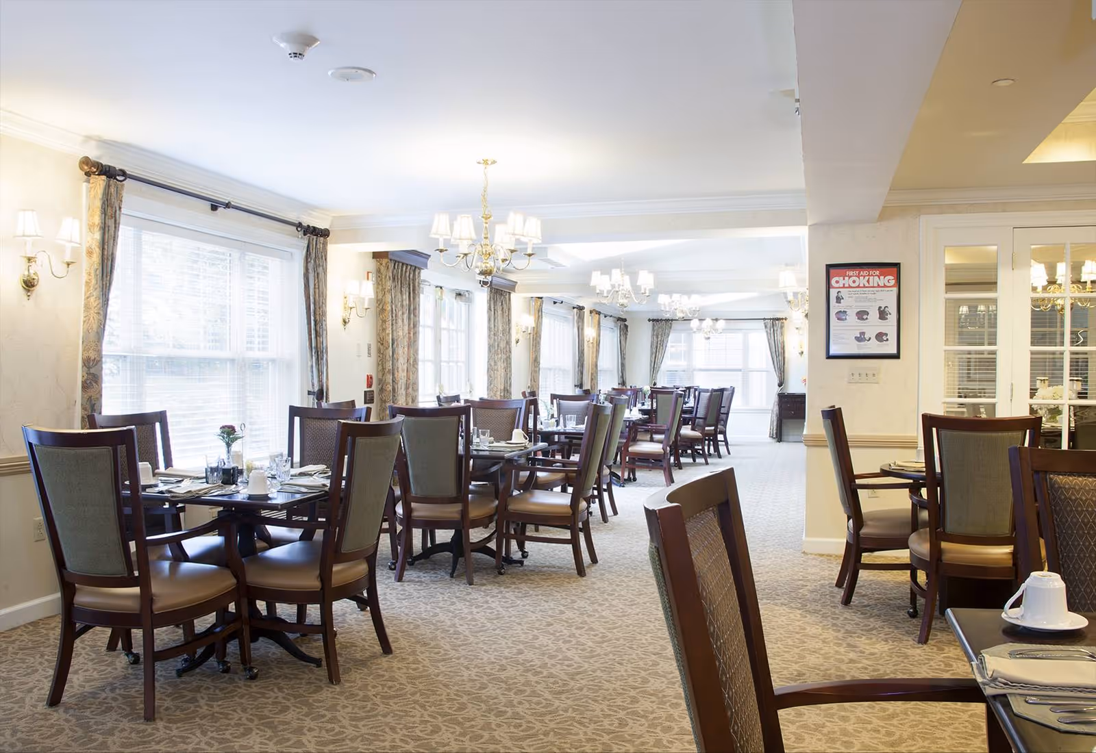 A bright and spacious dining room with multiple wooden tables and chairs arranged neatly. The room features large windows with curtains allowing natural light to fill the space. Elegant chandeliers and wall sconces provide additional lighting. Tables are set with plates, cups, and utensils, ready for dining.