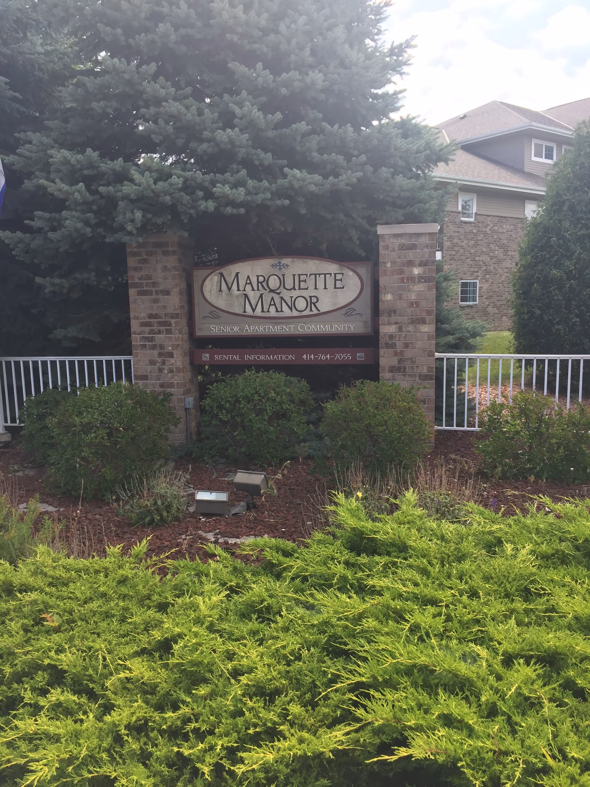 Outdoor view of the entrance sign for Marquette Manor Senior Apartment Community, surrounded by green bushes and trees, with a brick building visible in the background.