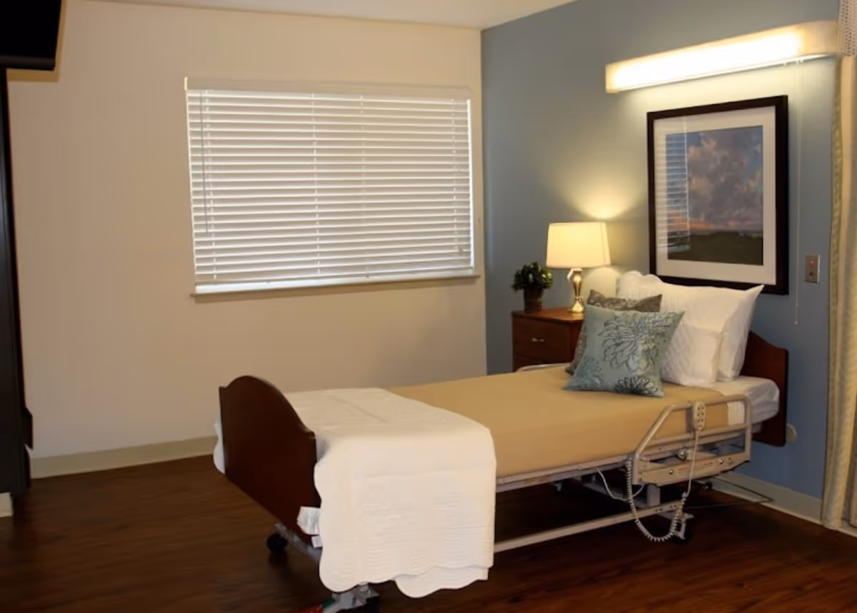 A tidy senior bedroom with an adjustable hospital bed, bedside table with lamp, framed artwork, and window blinds.
