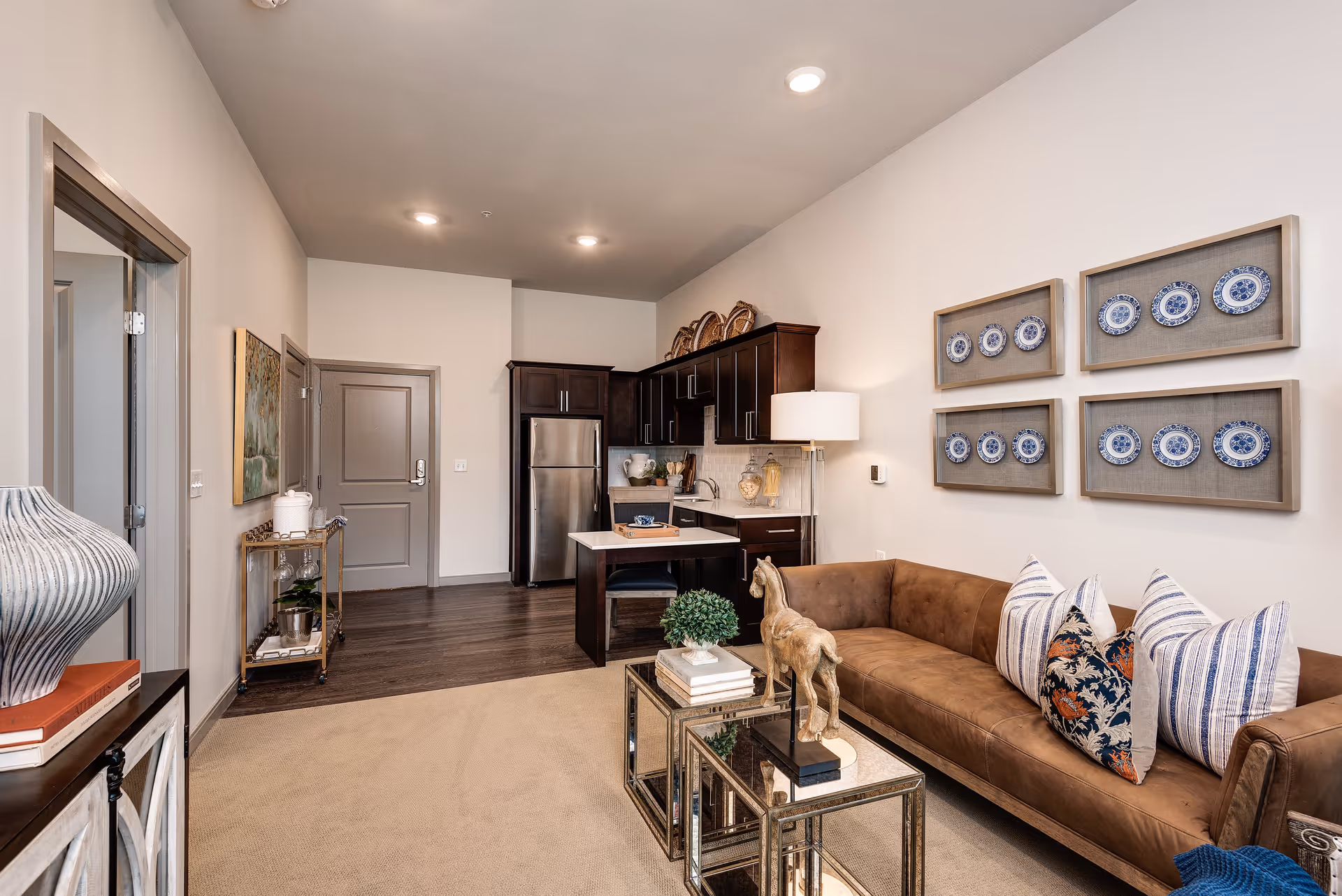 Interior view of a senior living facility apartment featuring a small kitchen with dark wood cabinets, stainless steel refrigerator, and a small island with a chair. Adjacent to the kitchen is a living area with a brown leather sofa adorned with patterned pillows, a glass coffee table with decorative items, and framed blue and white plates on the wall. The room has beige carpeting and wood flooring near the kitchen, with neutral-colored walls and ceiling lights.
