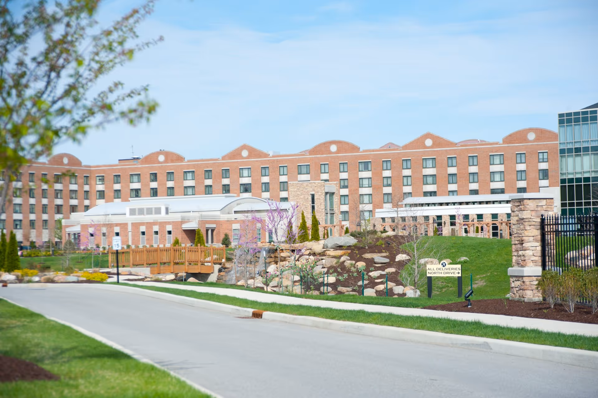 Front exterior of a large multi-story brick senior living building with a landscaped entrance, driveway, and stone features.
