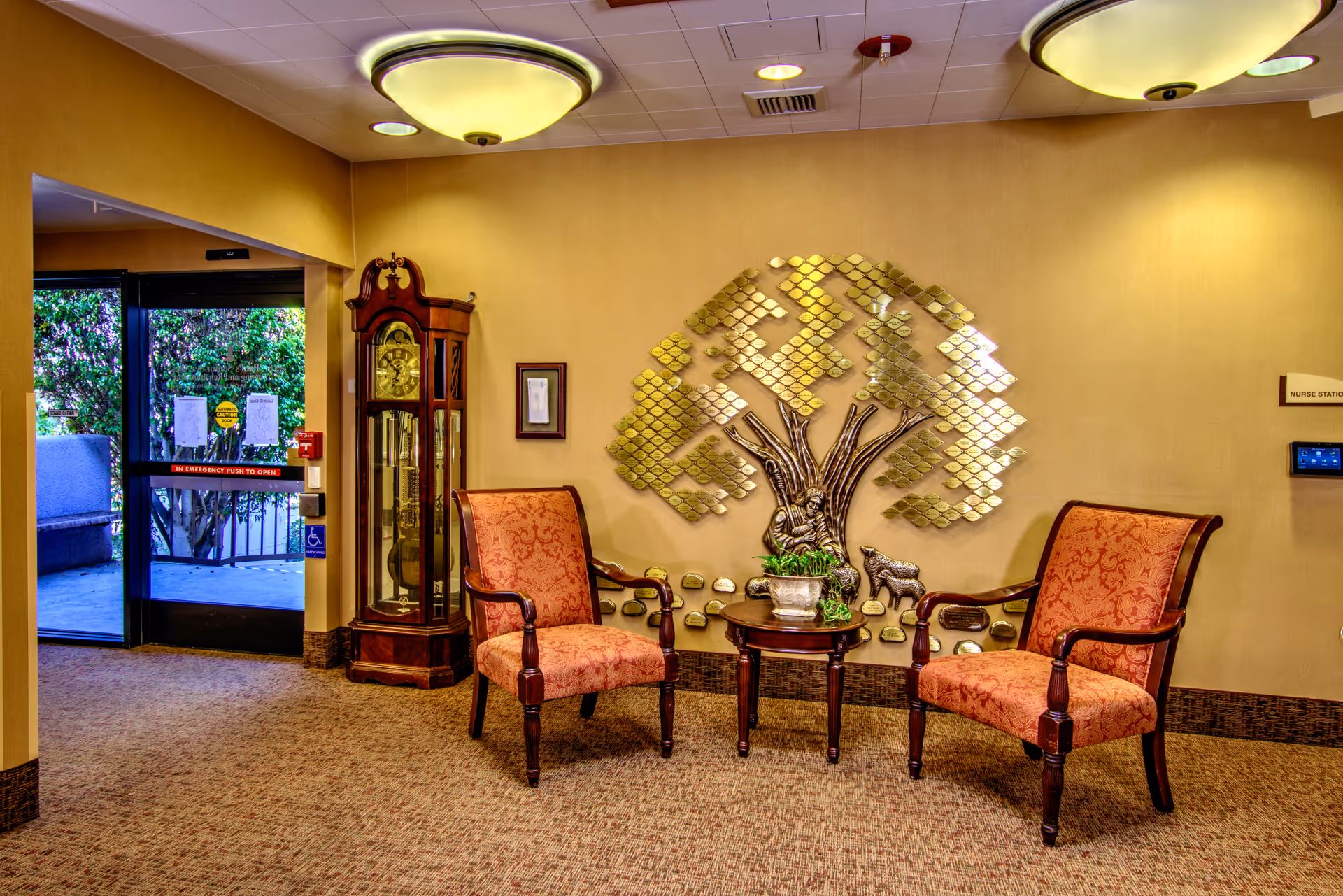 A cozy seating area in a nursing facility with two upholstered armchairs and a small round wooden table between them holding a potted plant. Behind the chairs is a decorative metallic wall art piece shaped like a tree with animals. To the left is a tall grandfather clock and an exit door leading outside. The walls are painted beige and the floor is carpeted.