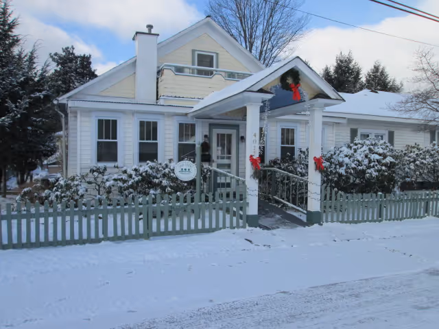 A white house with a covered entrance decorated with red bows and a wreath, surrounded by a green picket fence. Snow covers the ground, bushes, and roof, with trees in the background under a partly cloudy sky.