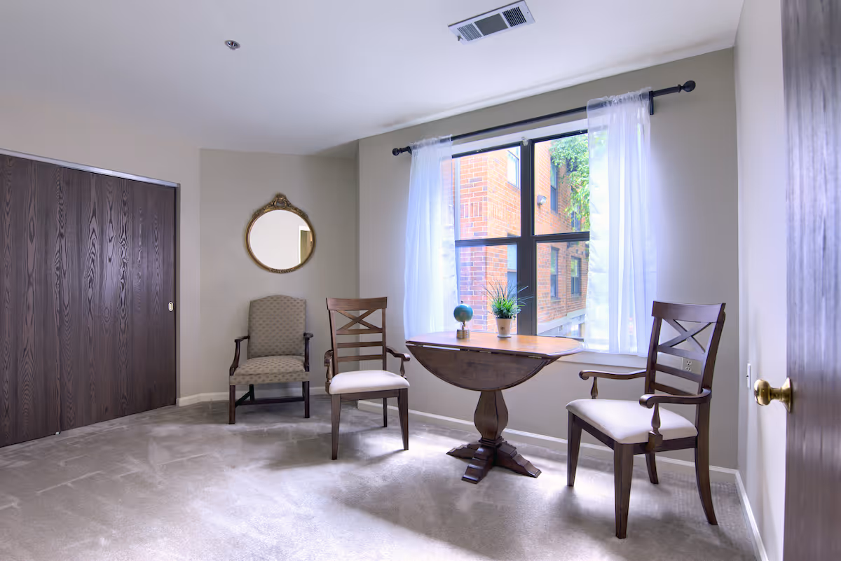 A small room with beige walls and carpeted floor featuring a wooden drop-leaf table with a small globe and a potted plant on top. There are three wooden chairs with cushioned seats around the table. A large window with sheer white curtains lets in natural light, showing a brick building outside. A round decorative mirror hangs on the wall above one of the chairs, and there is a dark wooden sliding closet door on the left side of the room.