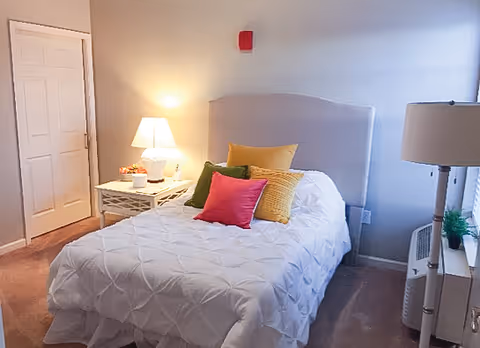 A cozy bedroom with a neatly made bed featuring a white quilt and colorful pillows in yellow, green, and pink. There is a white nightstand with a lamp and decorative items on the left side of the bed. A floor lamp and a window with blinds are on the right side, and a closed door is visible in the background.