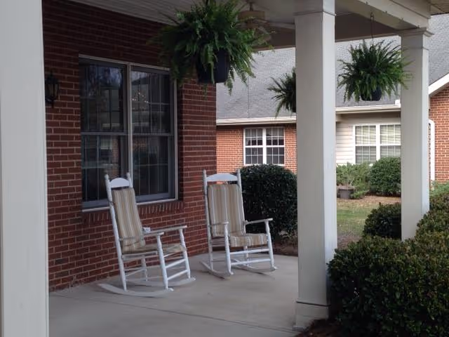 A covered porch area with two white rocking chairs with striped cushions. The porch has white pillars and hanging green ferns. In the background, there are brick and siding buildings with windows and bushes.