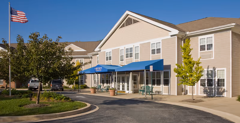 Exterior view of Lighthouse Senior Living at Hopkins Creek building with beige siding, white trim, and a blue awning over the entrance. There are trees, a flagpole with an American flag, and a curved driveway in front of the building under a clear blue sky.