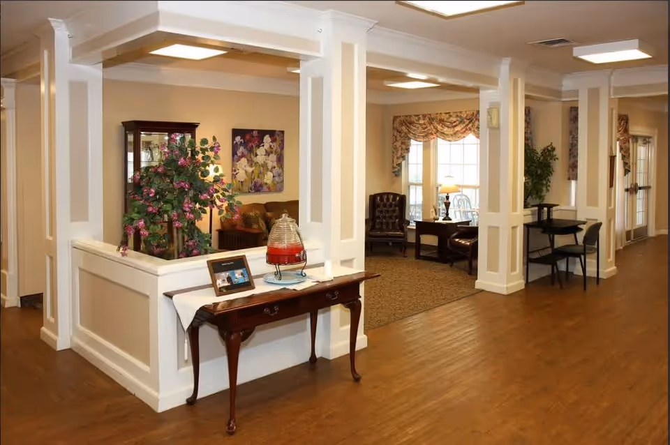 Interior view of a senior living facility lounge area with wooden flooring, white paneled columns, a small wooden table with a decorative drink dispenser and framed photo, a large plant, and seating areas with armchairs and side tables near windows with floral curtains.