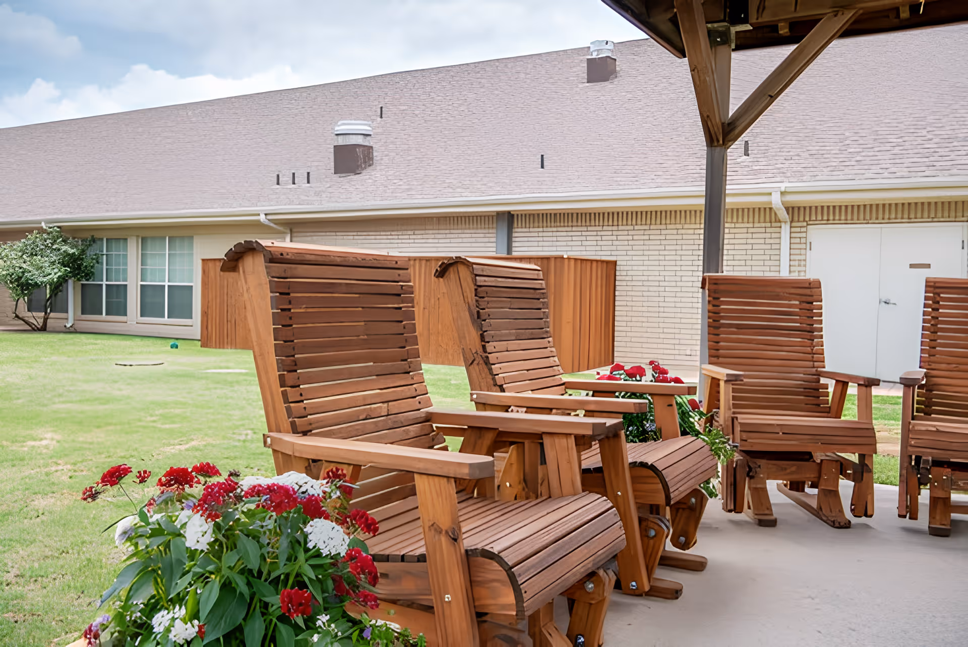 Outdoor covered patio area with wooden rocking chairs arranged in a semi-circle, surrounded by green grass and flower beds with red and white flowers. A beige brick building with windows and a door is visible in the background under a partly cloudy sky.