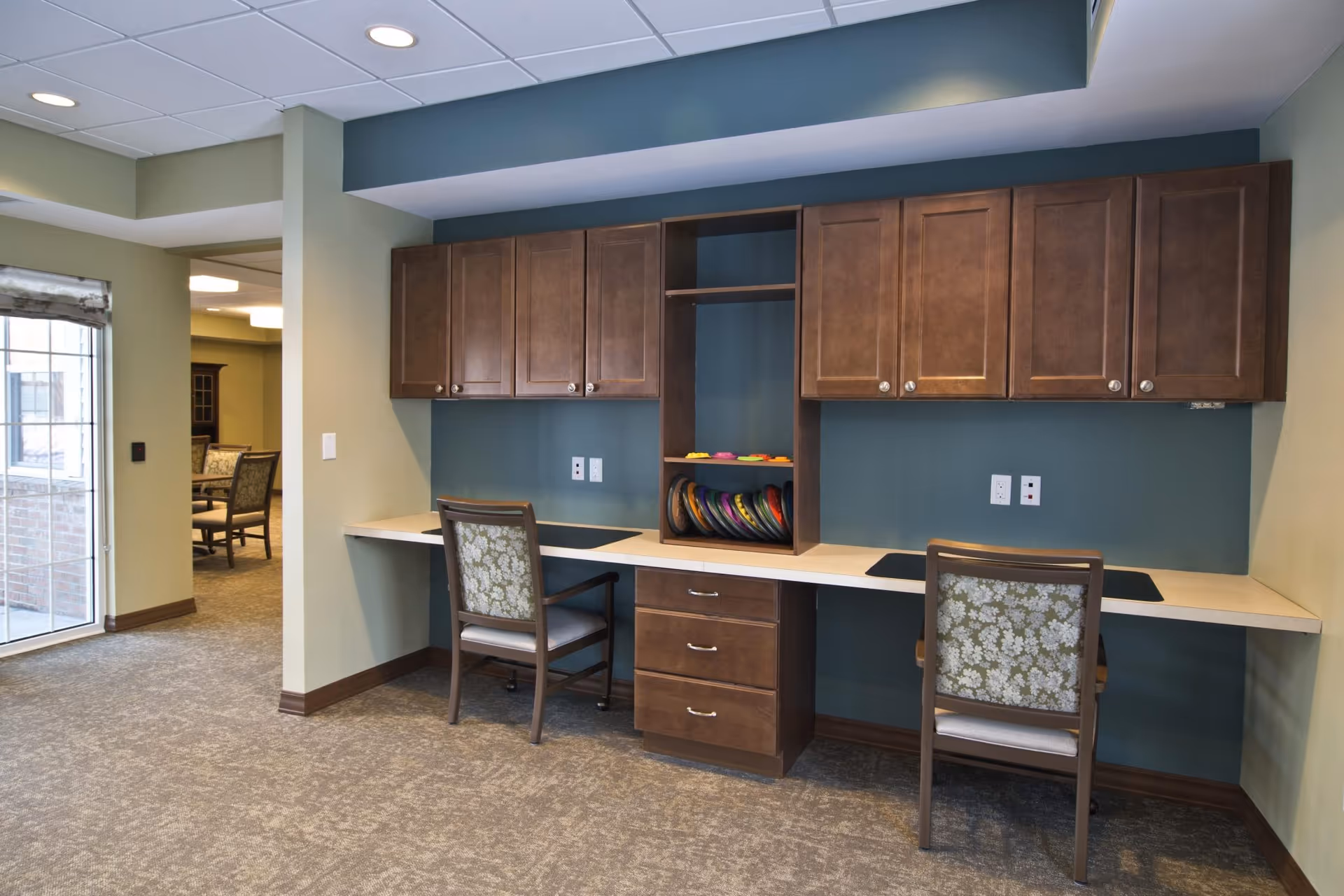 Built-in double workstation with two chairs, overhead cabinets and shelving in a carpeted common area.