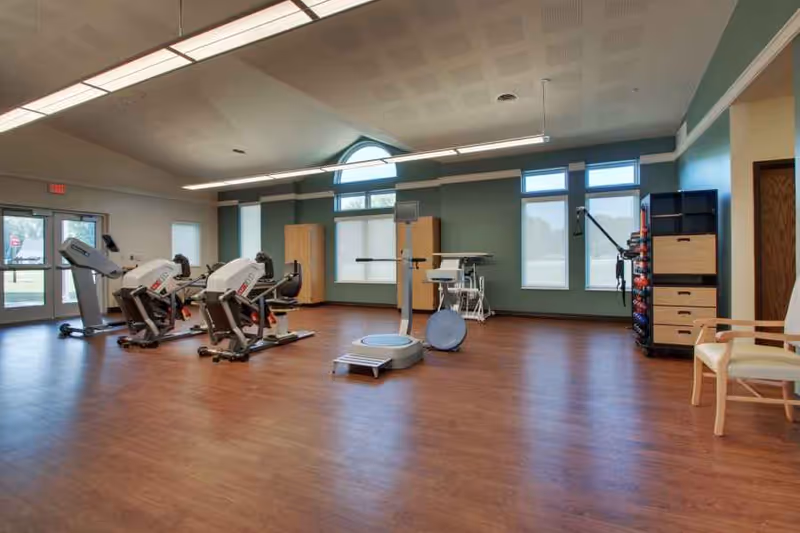 A spacious exercise room with wooden flooring and green walls, featuring several pieces of fitness equipment including recumbent bikes, a vibration plate machine, and a rack with various exercise balls and weights. There are large windows letting in natural light and a wooden chair in the corner.