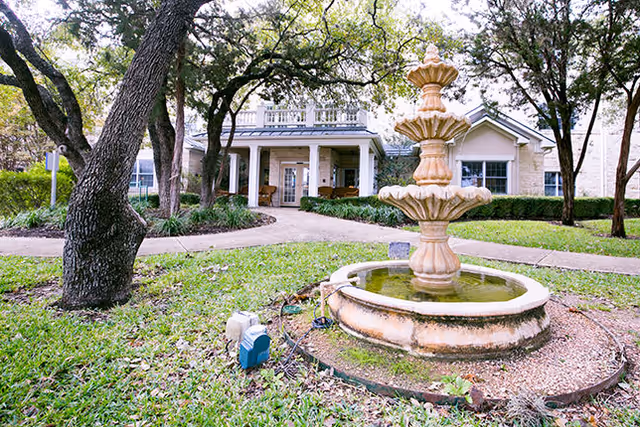 Three-tier stone fountain in a grassy courtyard in front of a single-story brick building entrance framed by trees.