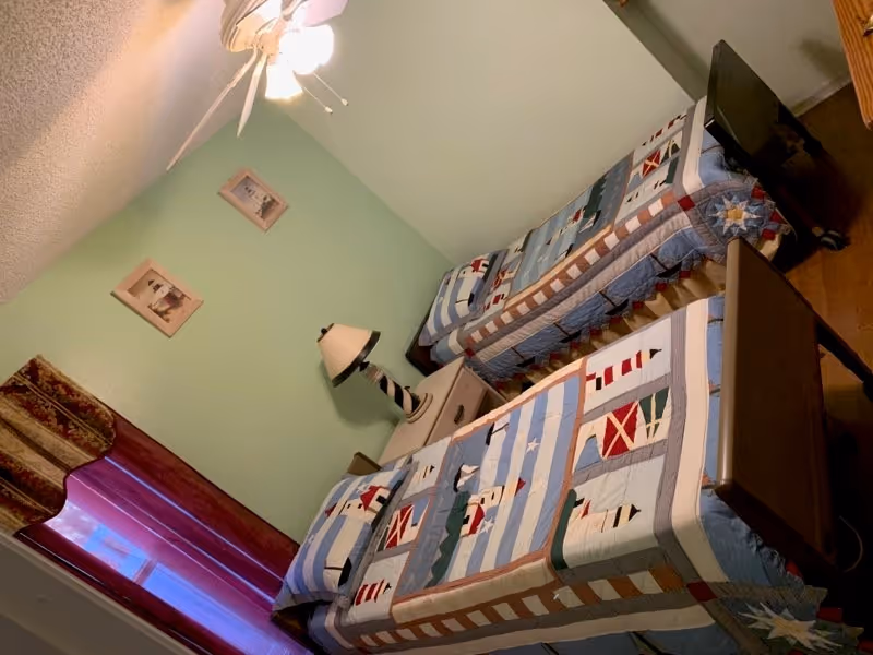 A bedroom with two twin beds covered in blue quilts featuring lighthouse and nautical designs. Between the beds is a nightstand with a lamp that has a striped base. The walls are painted light green, and there are two framed pictures hanging above the nightstand. A window with red curtains is visible on the left side of the room, and a ceiling fan with a light is on the ceiling.