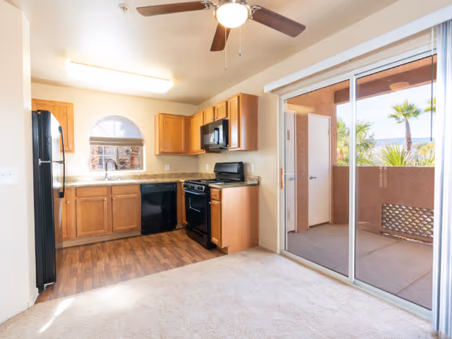 Interior view of a kitchen area with wooden cabinets, black appliances including a refrigerator, dishwasher, stove, and microwave. The kitchen has a small window above the sink and a ceiling fan with a light. Adjacent to the kitchen is a carpeted living space with large sliding glass doors leading to an outdoor patio with palm trees visible outside.
