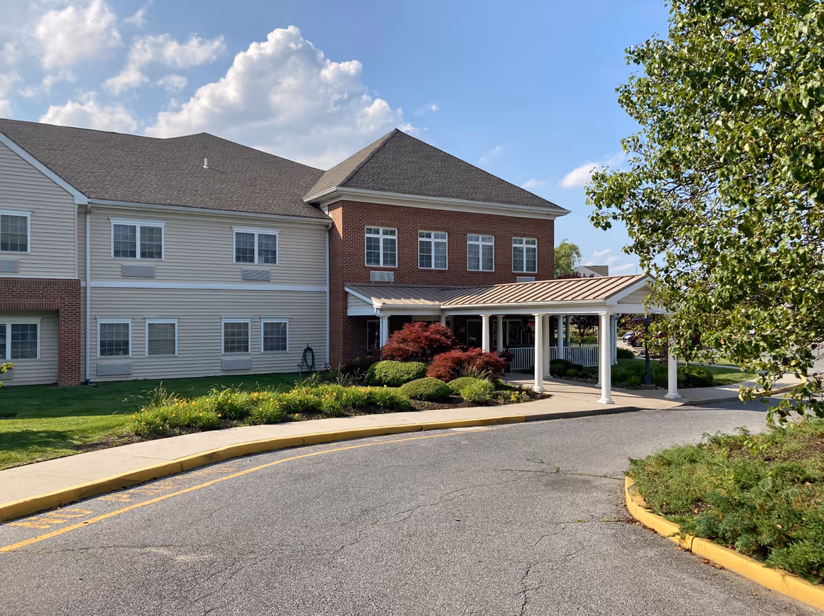 Front entrance of a two-story senior living building with a covered porte-cochere, landscaped shrubs, and a curved driveway.