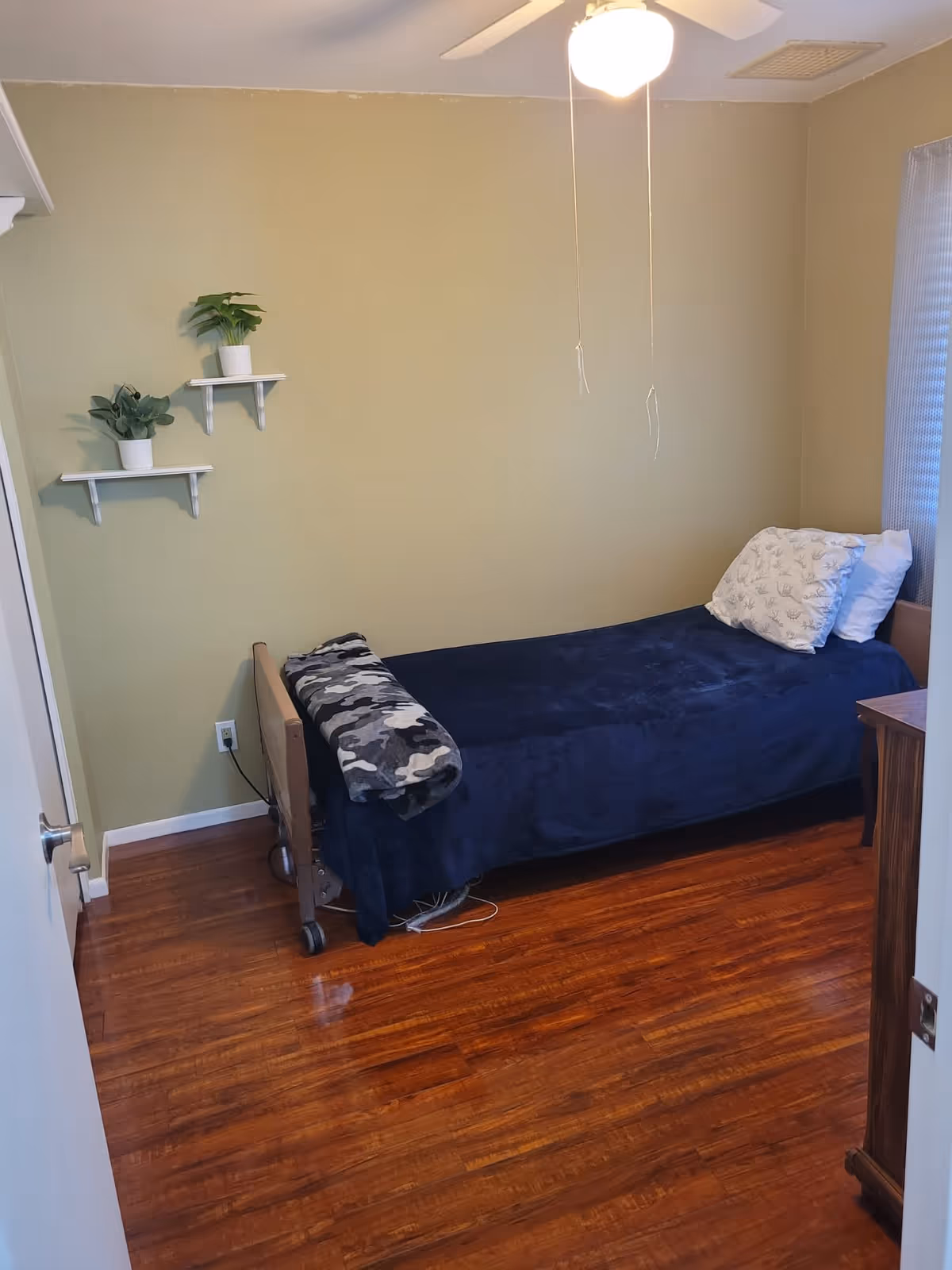 A small bedroom with a single bed covered in a dark blue blanket and a camouflage-patterned blanket folded at the foot. There are two pillows on the bed, one with a white cover and the other with a patterned cover. The room has wooden flooring, light green walls, and a ceiling fan with a light. Two small white shelves with potted plants are mounted on the wall above the bed. A window with a sheer curtain is visible on the right side.