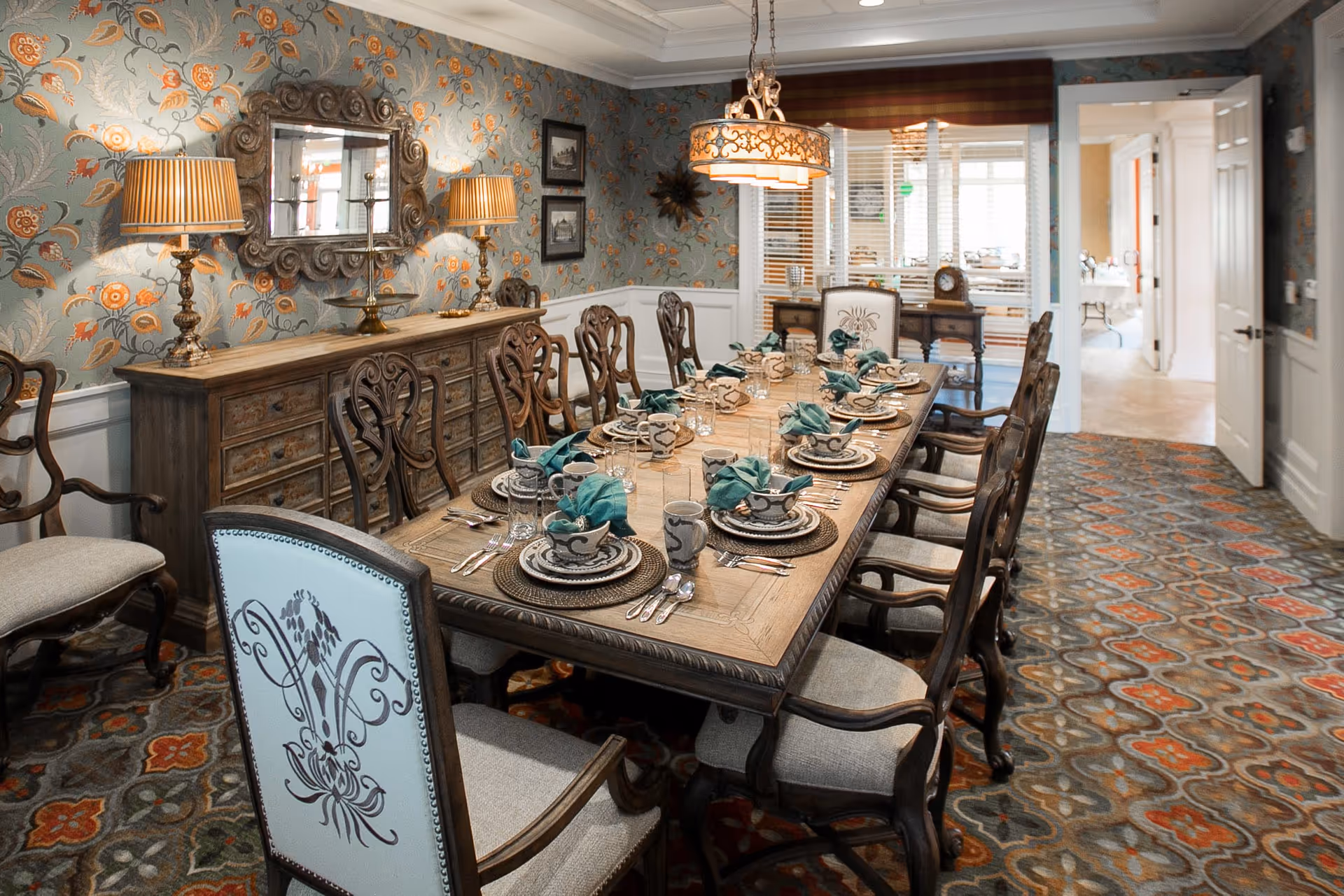 A formal dining room with a long wooden table set for twelve people with patterned plates, cups, and teal napkins. The room features ornate wooden chairs, a sideboard with two lamps and a decorative mirror above it, floral wallpaper, and a patterned carpet. A chandelier hangs above the table, and there is a doorway leading to another room in the background.