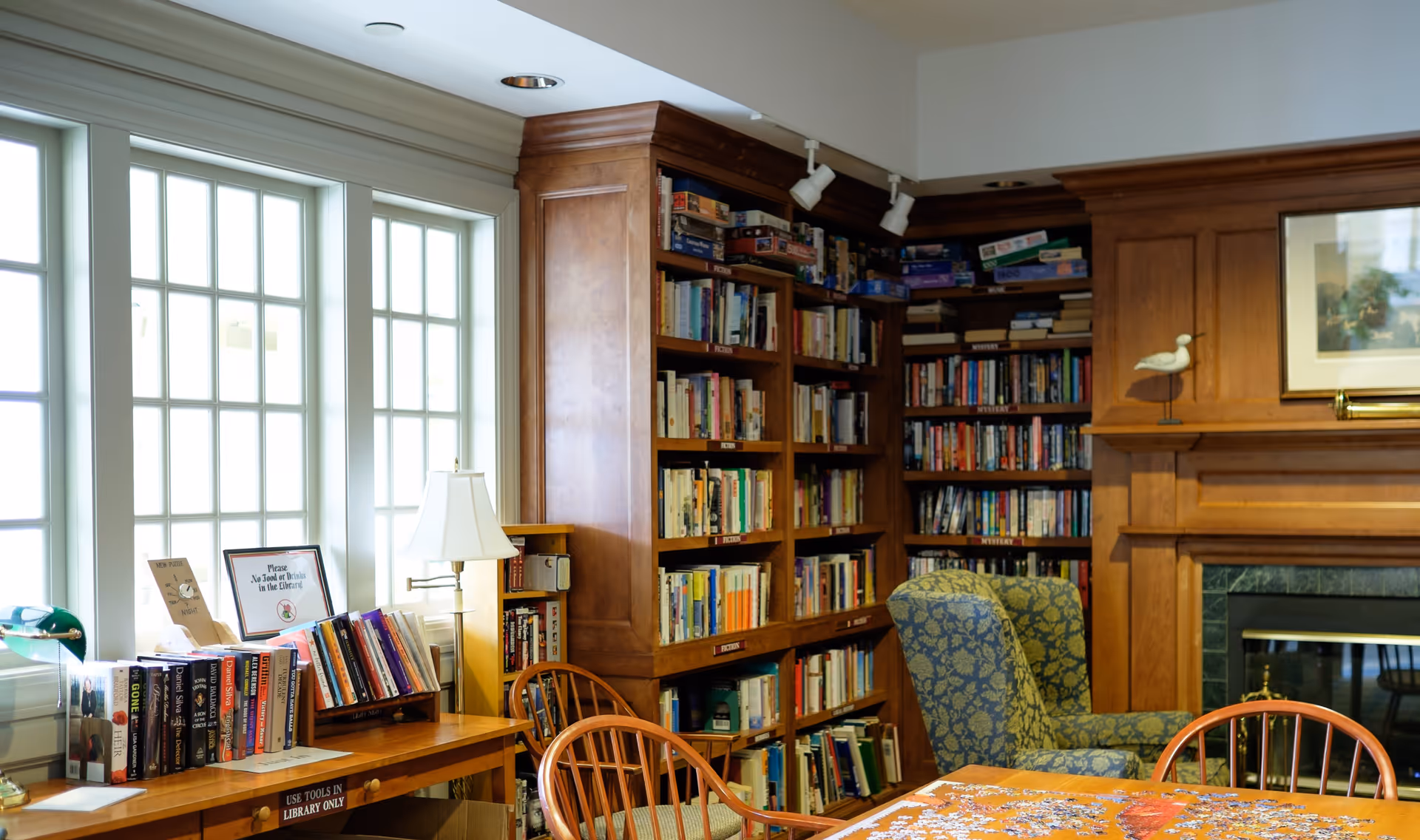 A cozy library room with large windows letting in natural light, wooden bookshelves filled with books and board games, a comfortable upholstered armchair, a wooden table with chairs, and a partially completed jigsaw puzzle on the table. There is also a lamp and framed artwork above a fireplace.