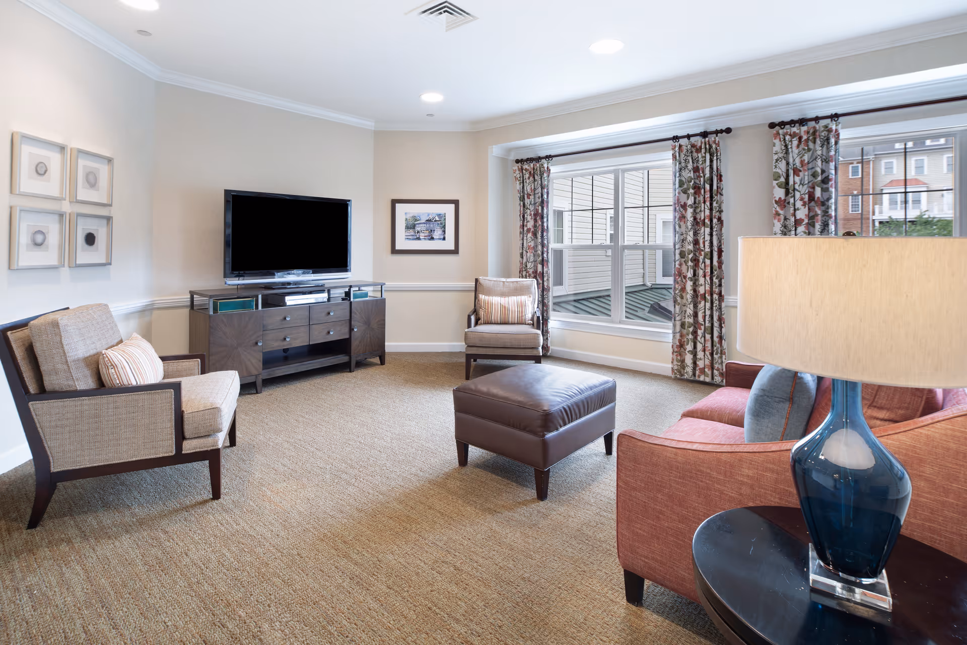 A bright and cozy living room with beige walls and carpet. The room features a flat-screen TV on a dark wooden console, two beige armchairs with striped cushions, a brown leather ottoman, and a coral-colored sofa with blue and coral pillows. A blue glass table lamp sits on a round dark wooden side table. Large windows with floral curtains allow natural light to fill the space.
