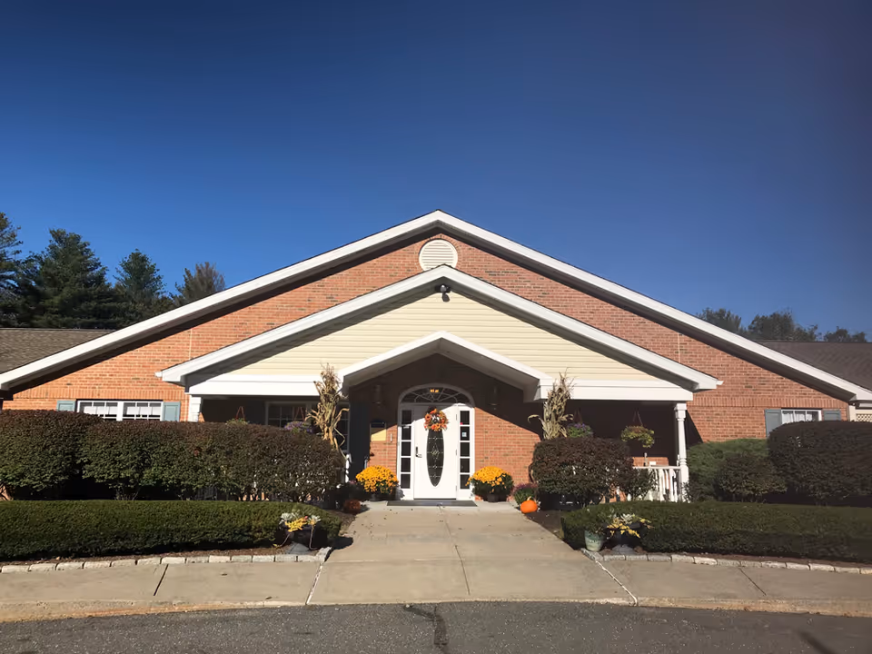 Front exterior view of a single-story brick building with a peaked roof and a covered entrance. The entrance door is white with glass panels and decorated with a fall-themed wreath. There are potted flowers and pumpkins on either side of the entrance, and neatly trimmed bushes line the walkway leading to the door. The sky is clear and blue.
