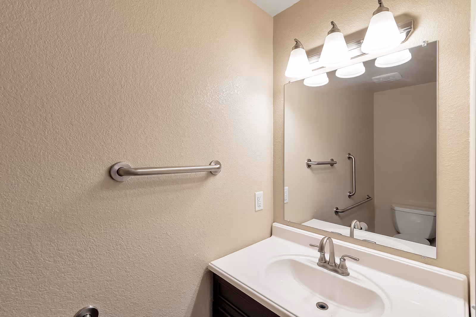 A clean bathroom with beige textured walls, a white sink with a silver faucet, a large rectangular mirror above the sink, and three light fixtures mounted above the mirror. A silver towel bar is mounted on the wall to the left, and a toilet with grab bars is visible in the reflection of the mirror.