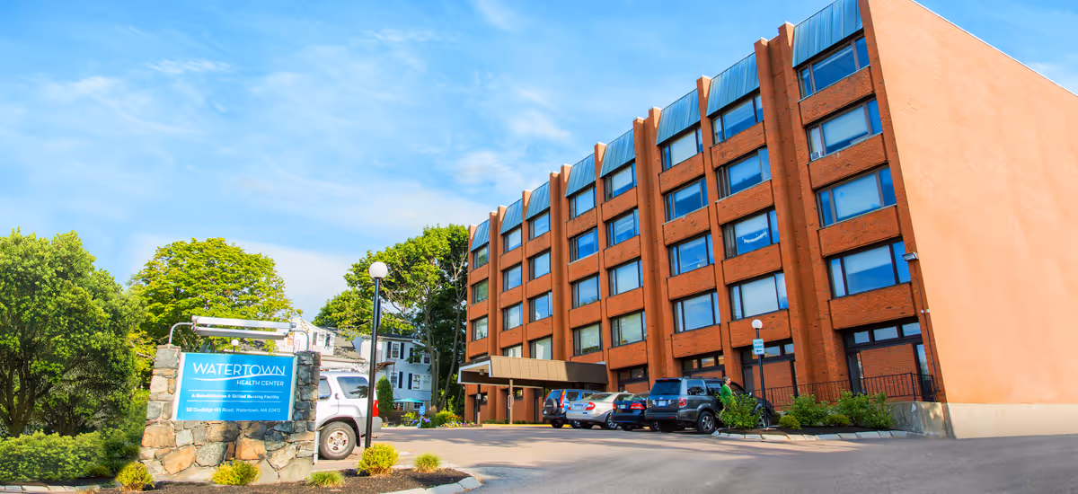 Brick building labeled Watertown Health Center with parking lot and green trees.