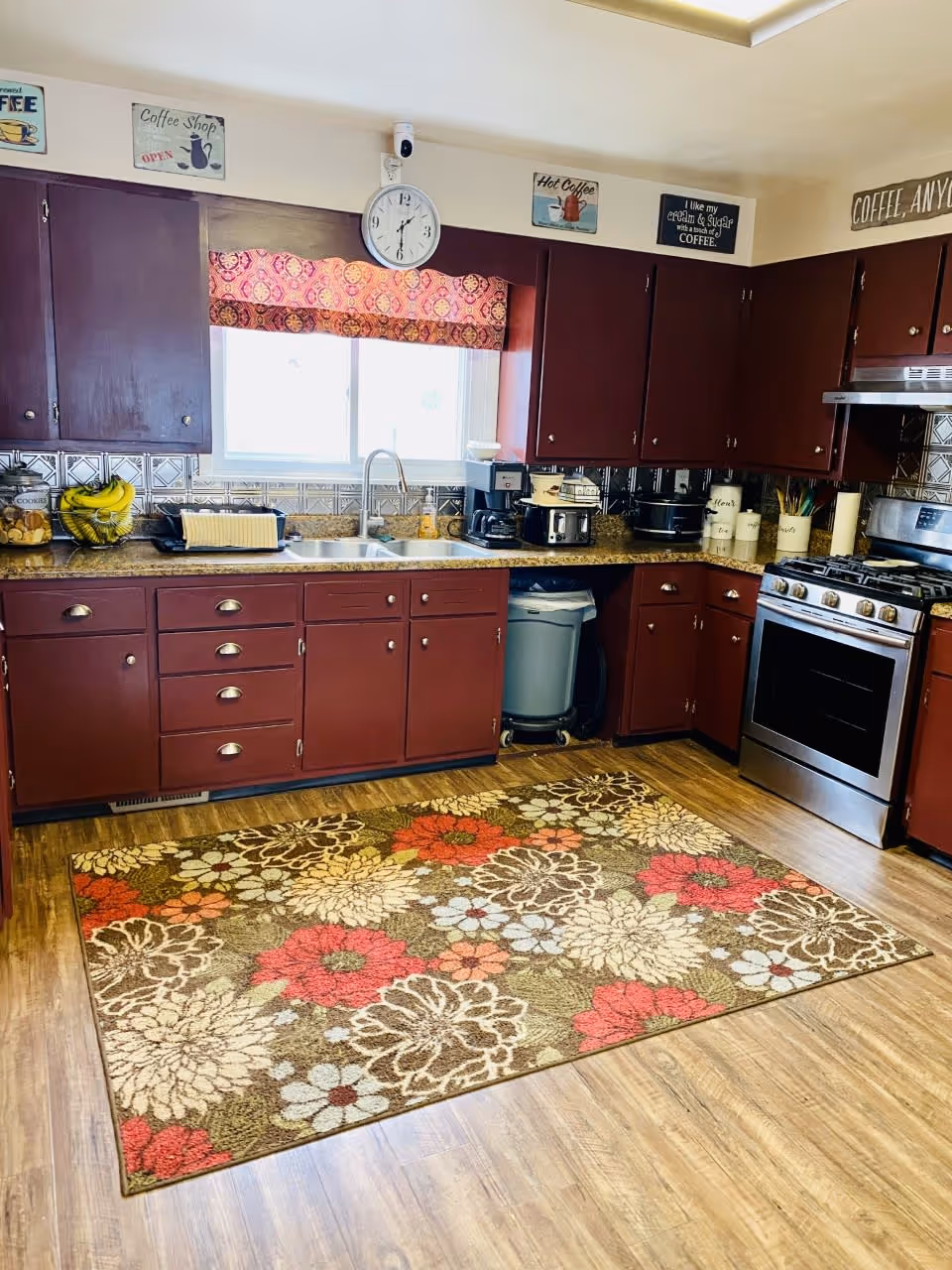 A kitchen with dark red cabinets, a stainless steel stove, and a double sink under a window with a patterned valance. The countertop holds various kitchen appliances including a coffee maker, toaster, and crockpots. The floor has a colorful floral area rug, and the walls are decorated with coffee-themed signs and a clock above the window.