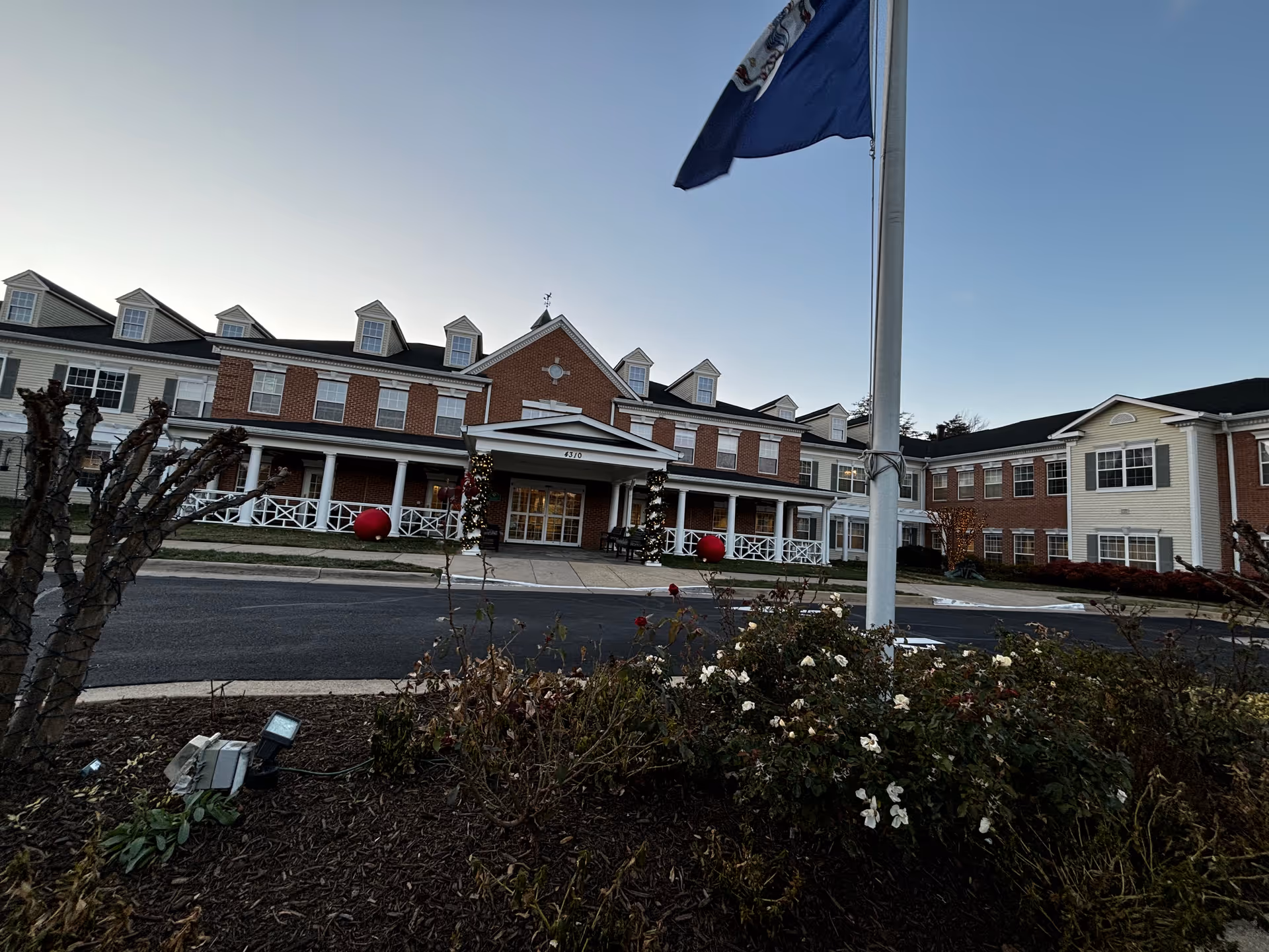 Exterior view of a senior living facility building with a brick and siding facade, multiple windows, and dormer windows on the roof. The entrance is decorated with holiday lights and large red ornaments. A flagpole with a flag is in the foreground, along with landscaped bushes and flowers.