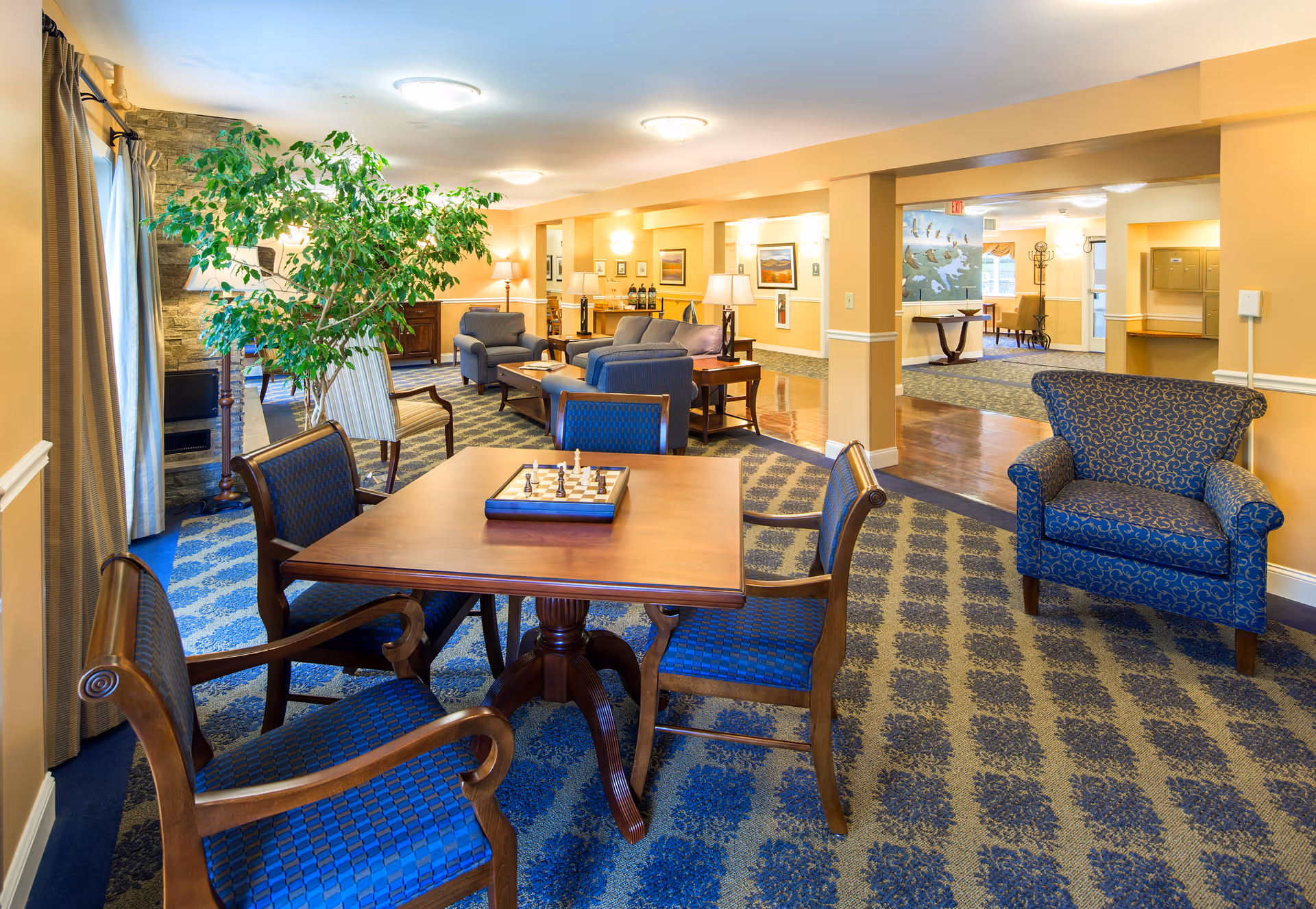 A spacious and well-lit common area in an assisted living facility featuring a wooden table with four blue cushioned chairs and a chessboard on top. The room has patterned blue carpeting, a large potted plant near a window with curtains, several armchairs and sofas arranged for conversation, warm yellow walls with framed artwork, and soft lighting from ceiling fixtures and table lamps.