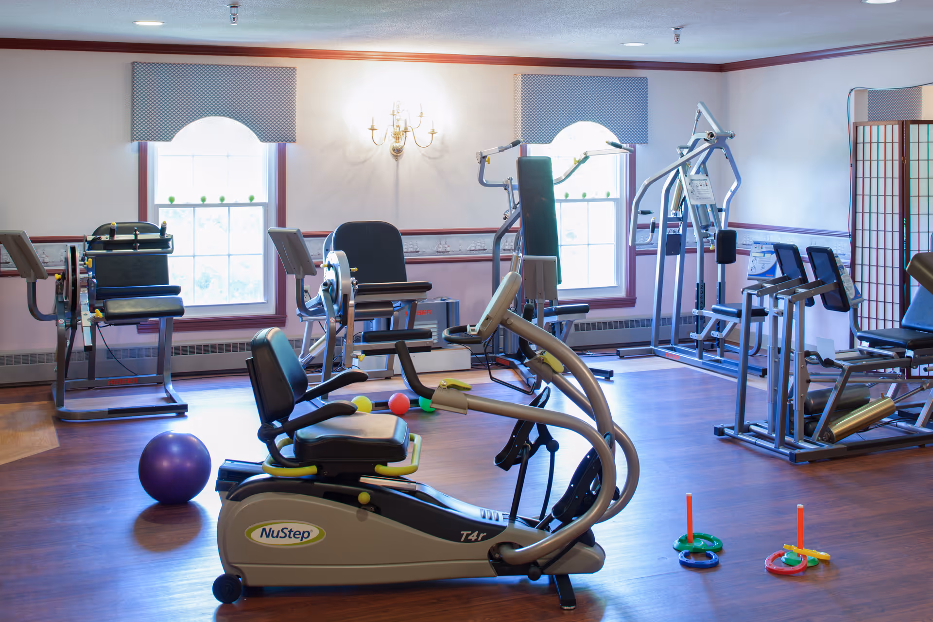 A well-lit exercise room with various fitness equipment including a NuStep T4r recumbent cross trainer, weight machines, and exercise balls. The room has wooden flooring, two large windows with valances, and a wall-mounted light fixture. There are also colorful ring toss game pieces on the floor.