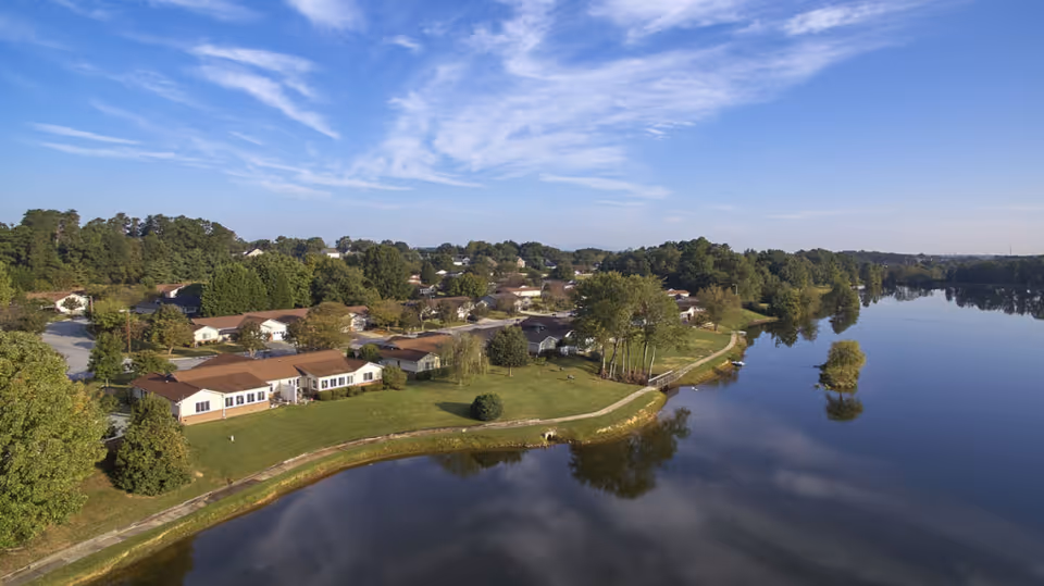 Aerial view of Rolling Green Village, showing a cluster of single-story residential buildings with brown roofs surrounded by green lawns and trees. The village is adjacent to a calm body of water reflecting the blue sky with scattered clouds. A walking path runs along the water's edge.