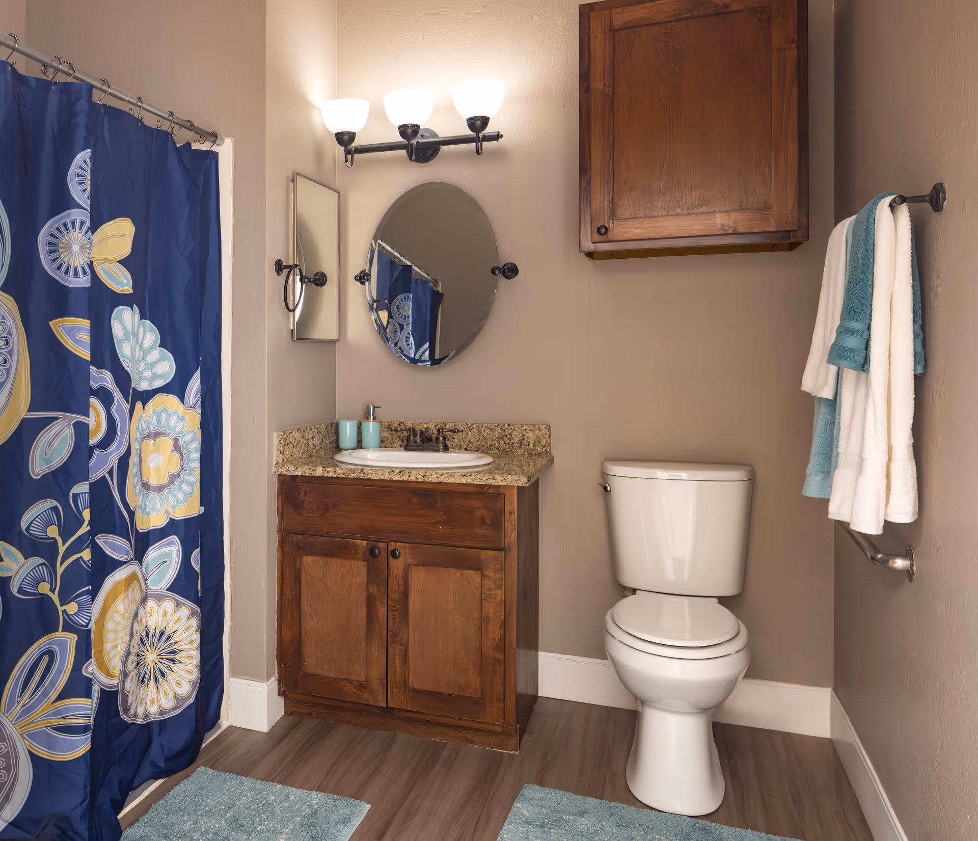 A bathroom featuring a white toilet, a wooden vanity with a granite countertop and a white sink, an oval mirror above the sink, a wall-mounted wooden cabinet, a towel rack with white and teal towels, and a shower with a blue floral-patterned curtain. The floor has wood-like flooring with two blue bath mats.
